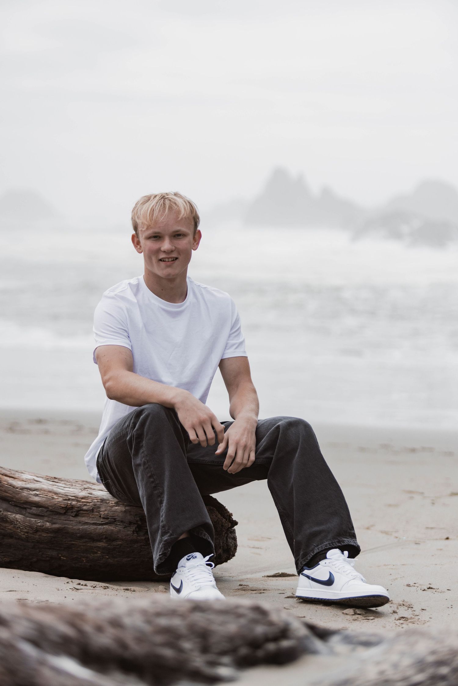 A person in a white t-shirt sits casually on driftwood at a misty beach wearing Nike sneakers.
