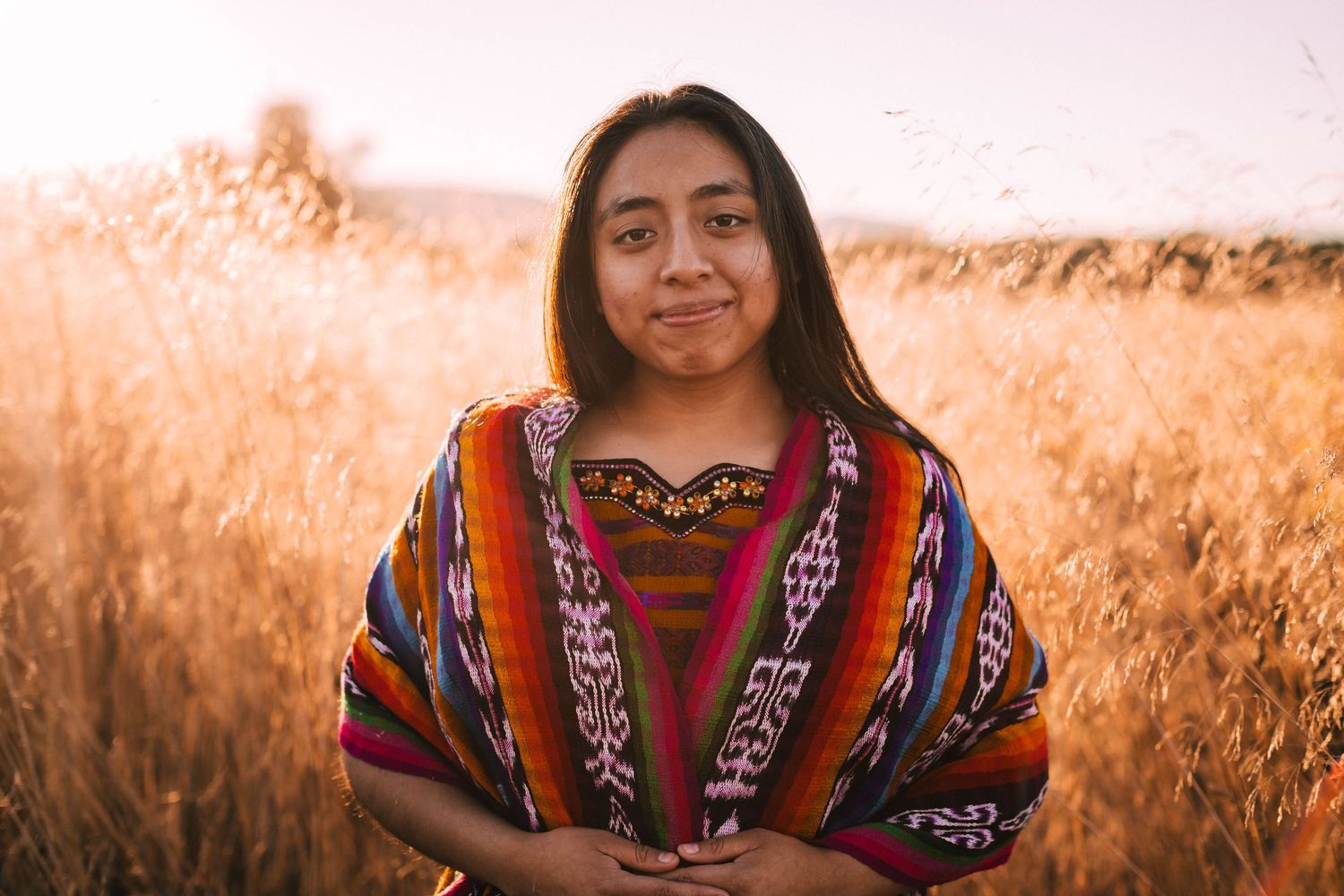 A person wearing traditional indigenous clothing poses in a golden wheat field at sunset.