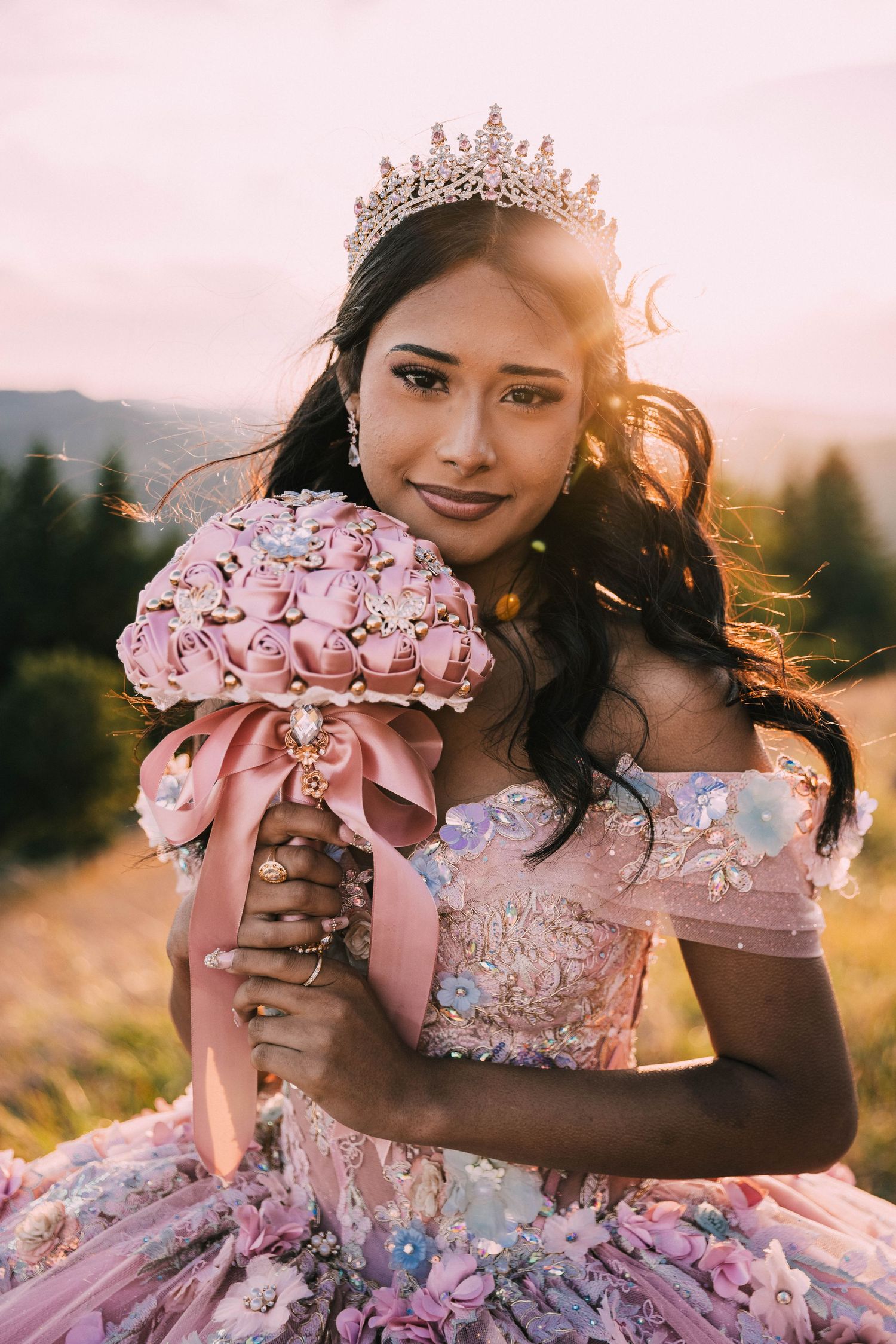 A person wearing a pink princess gown and crown holds a matching bouquet against a mountain landscape at golden hour.