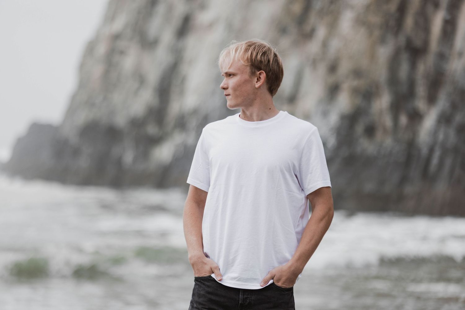 Person in white t-shirt and jeans stands against rocky coastal backdrop near ocean waves.