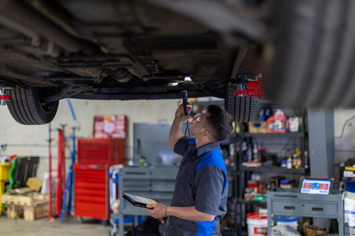 Auto mechanic inspects car suspension components in a professional repair shop with red tool cabinets visible.