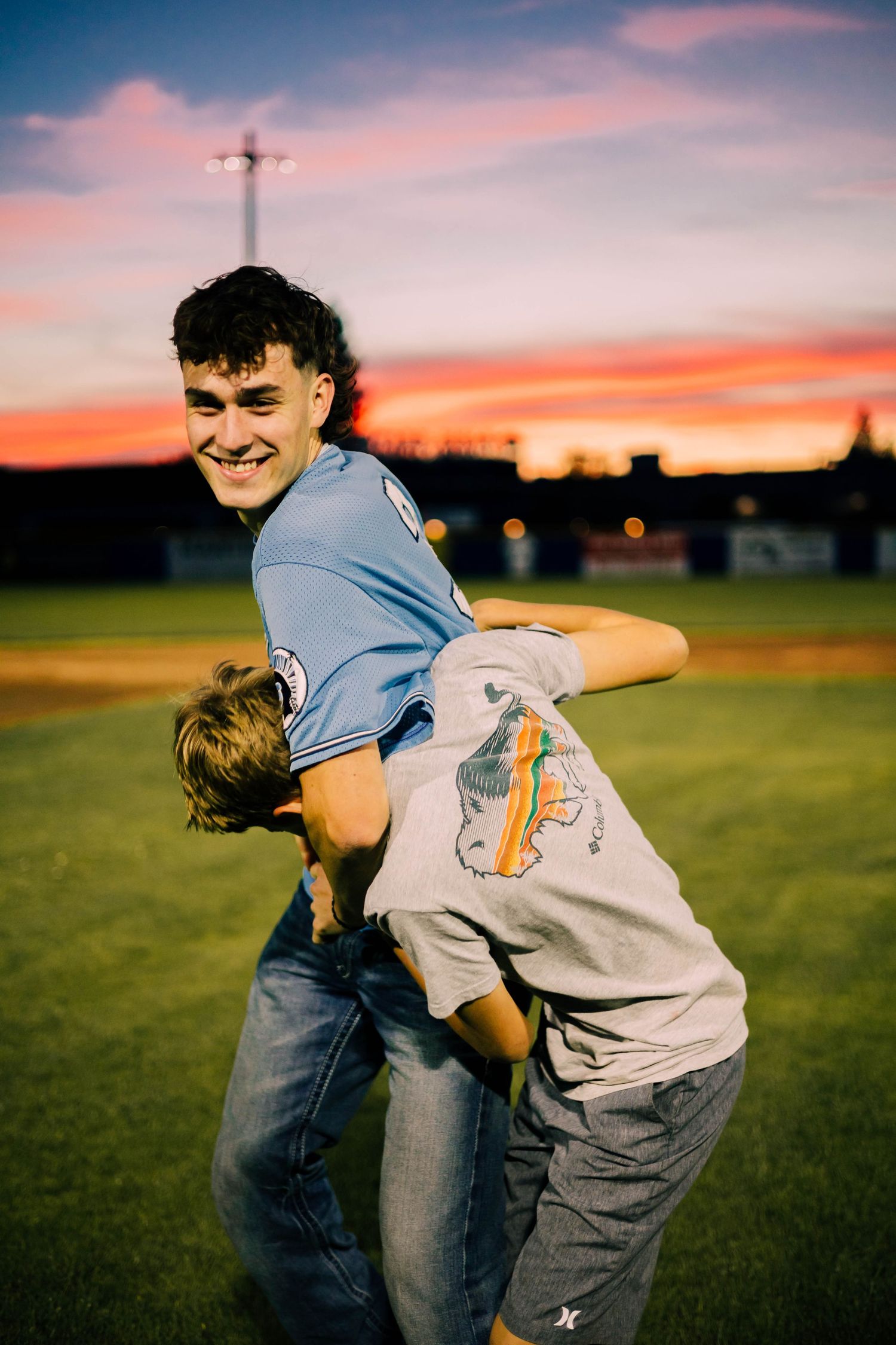 Two people in casual attire embrace playfully on a grassy field at sunset with vibrant pink and orange sky.