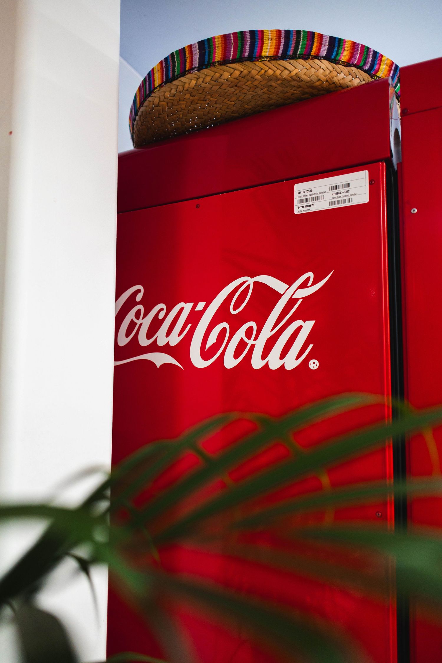 A vintage red Coca-Cola vending machine photographed through green plant leaves creating a shadowy overlay effect.