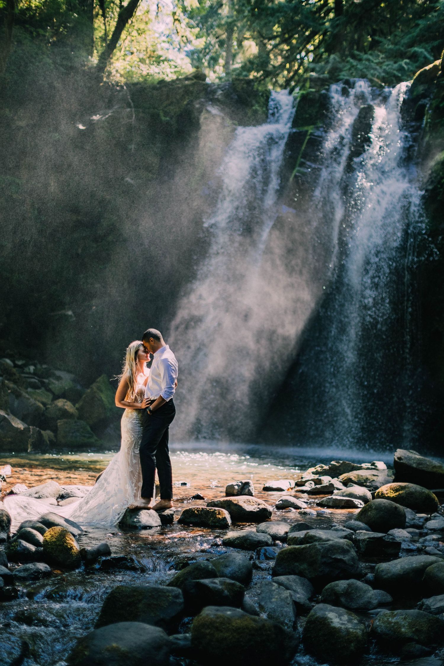 Dramatic waterfall creates misty backdrop for romantic wedding photos on rocky shore.