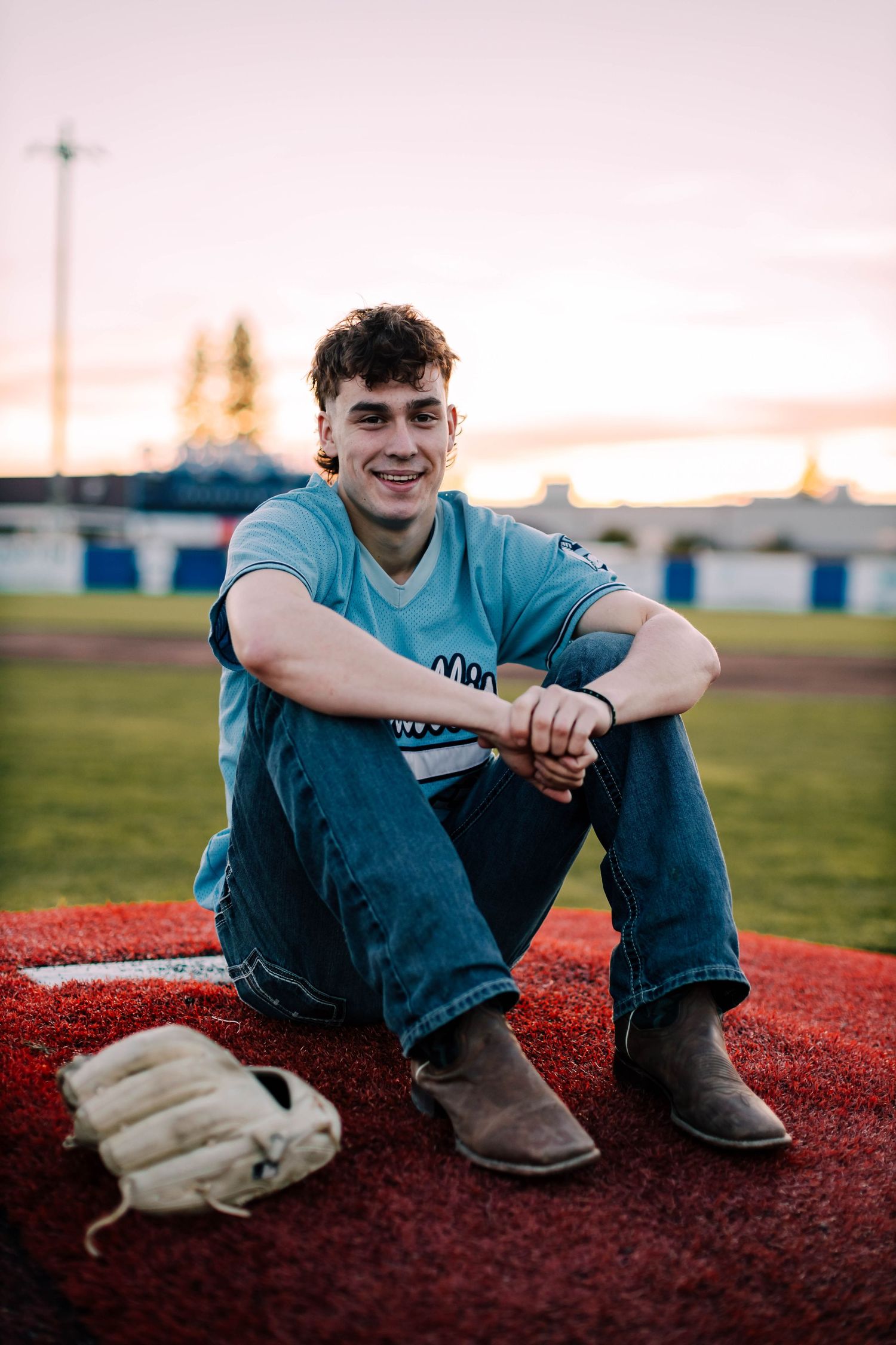Baseball player sits on red track surface with baseball glove beside them at sunset.