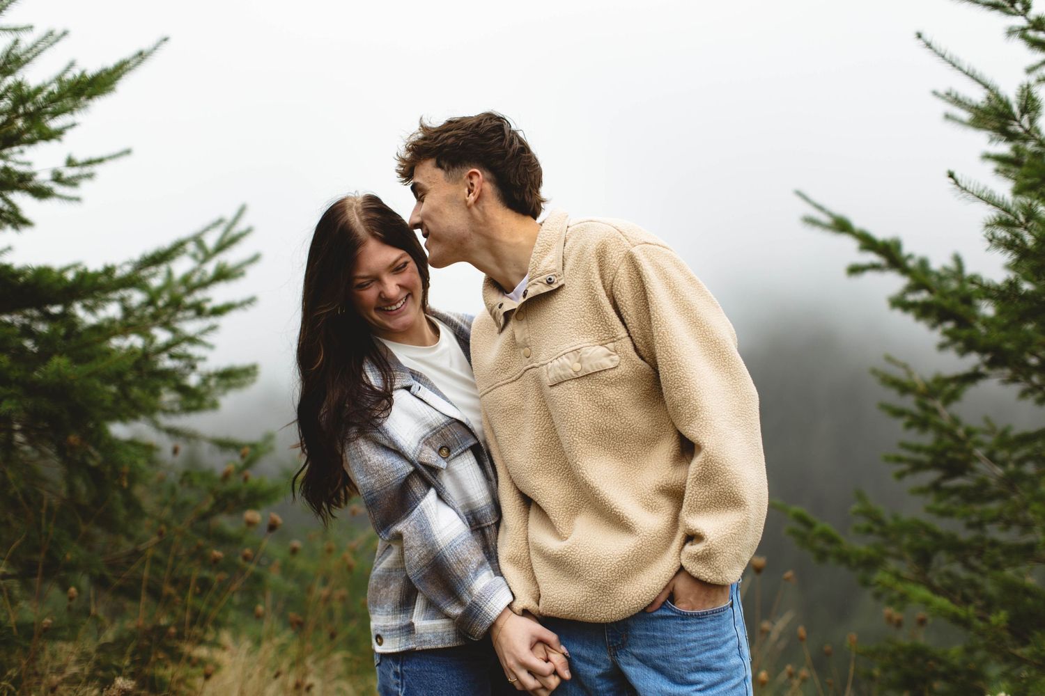 A couple in casual attire embraces while standing in a misty mountain forest setting.