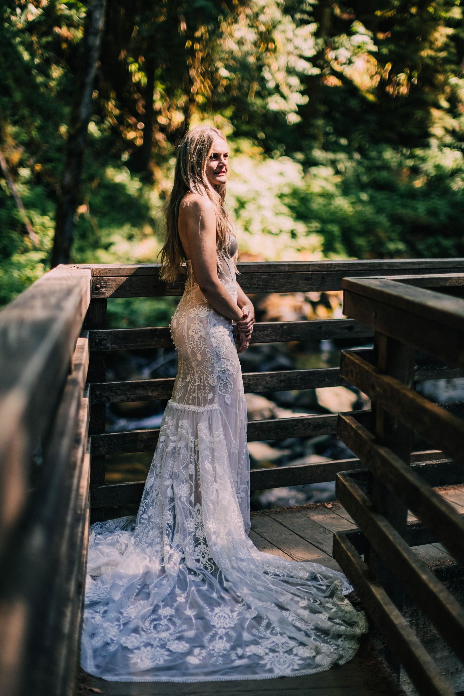 Elegant lace wedding dress trails across wooden bridge deck in natural forest lighting.