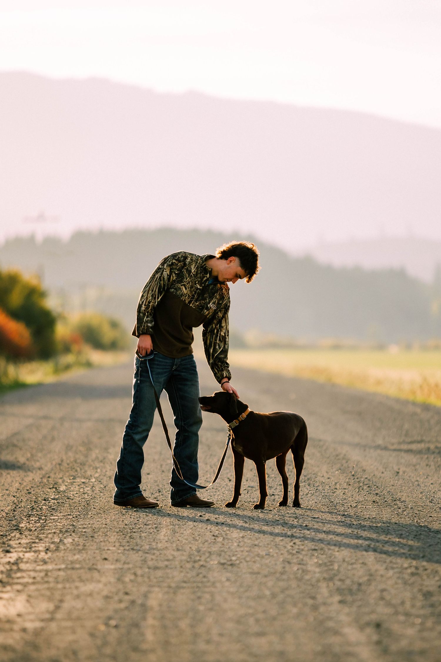 Person in camouflage shirt and jeans stands with their chocolate lab on a rural dirt road at sunset.
