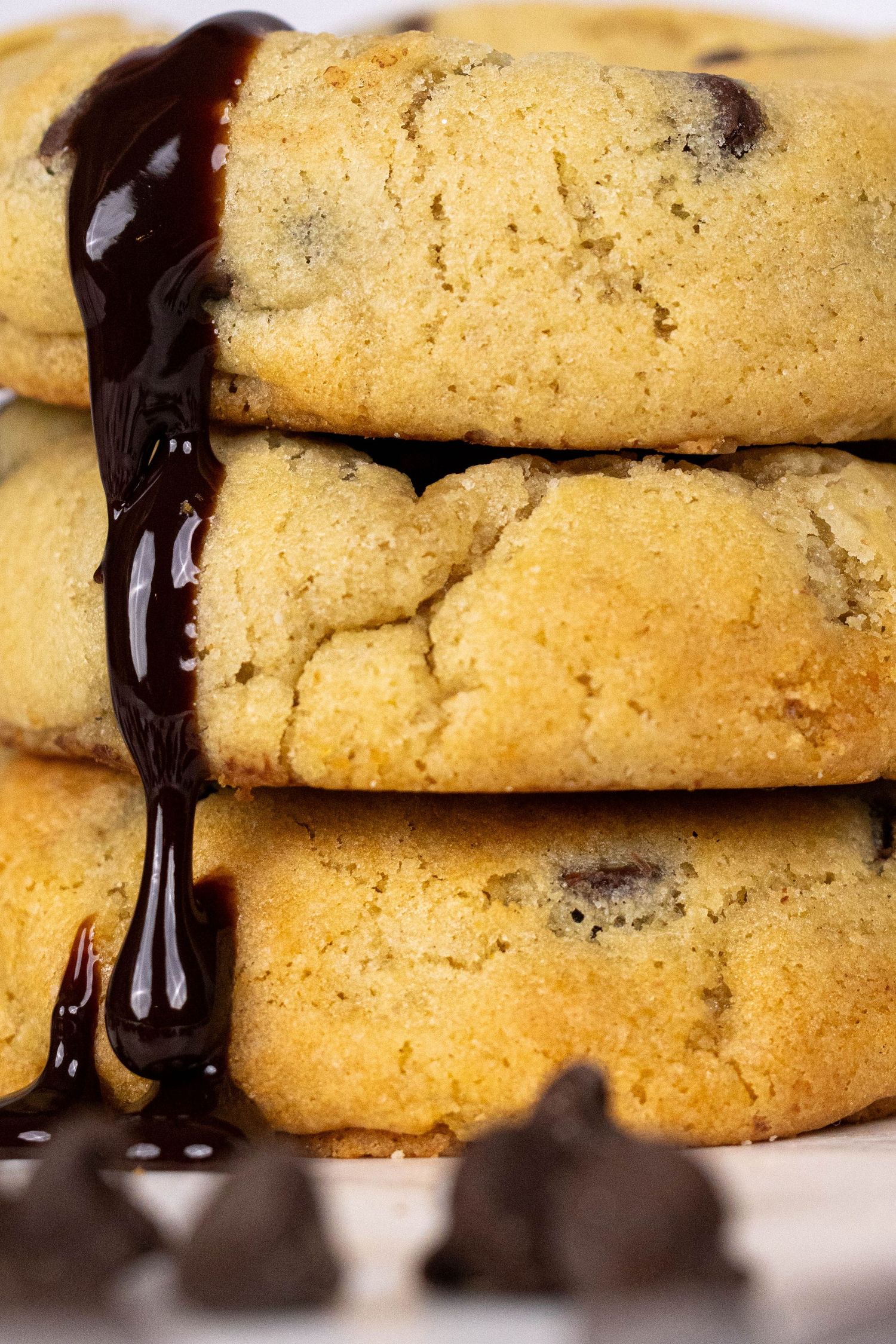 Fresh chocolate chip cookies stacked on a white surface with dark chocolate drizzle running down the sides.