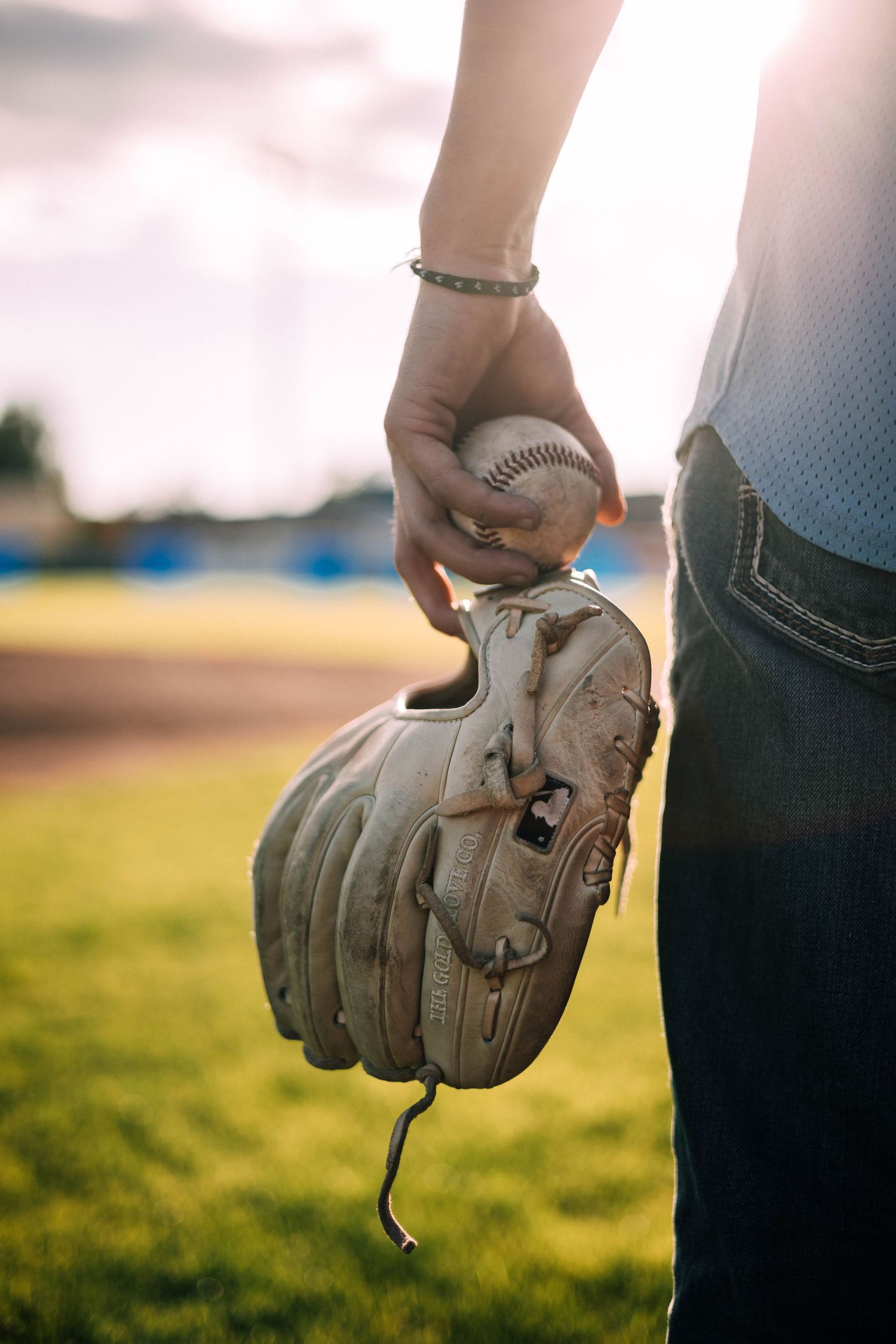 Hand wearing a baseball glove reaches down to pick up a baseball on a sunny field.