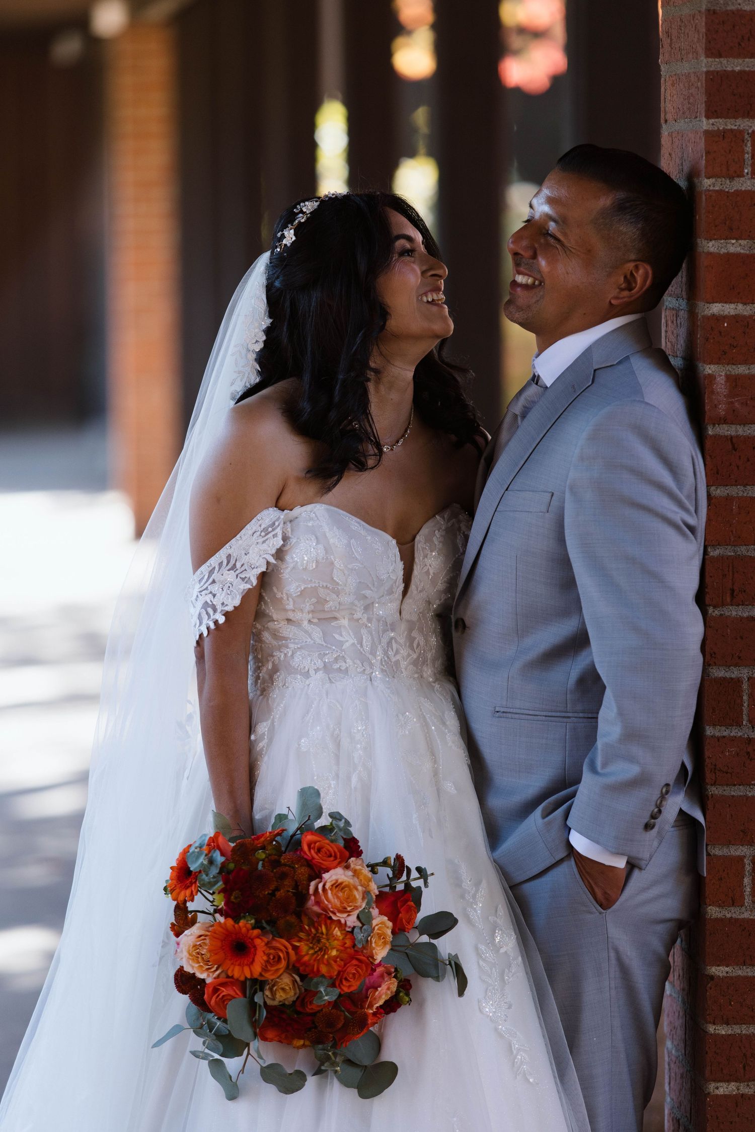 Newlywed couple shares intimate moments against brick wall while holding vibrant orange and red floral bouquet.