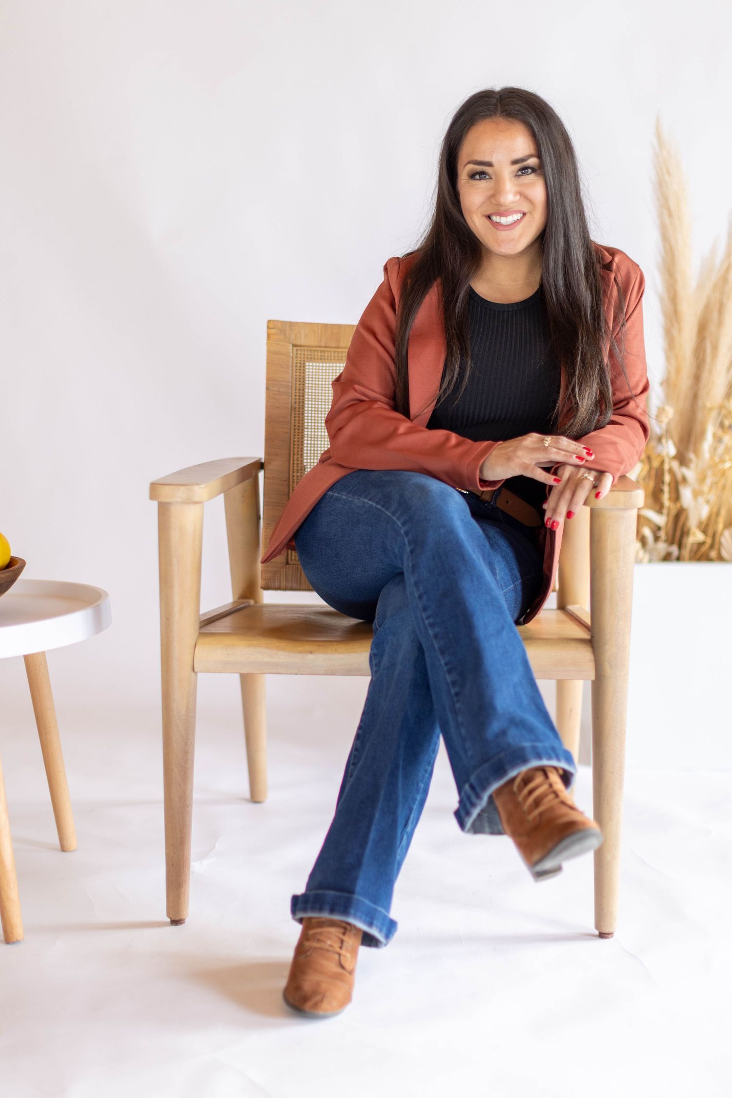 Person in black sweater and rust cardigan poses casually on wooden chair with pampas grass decor in white studio.