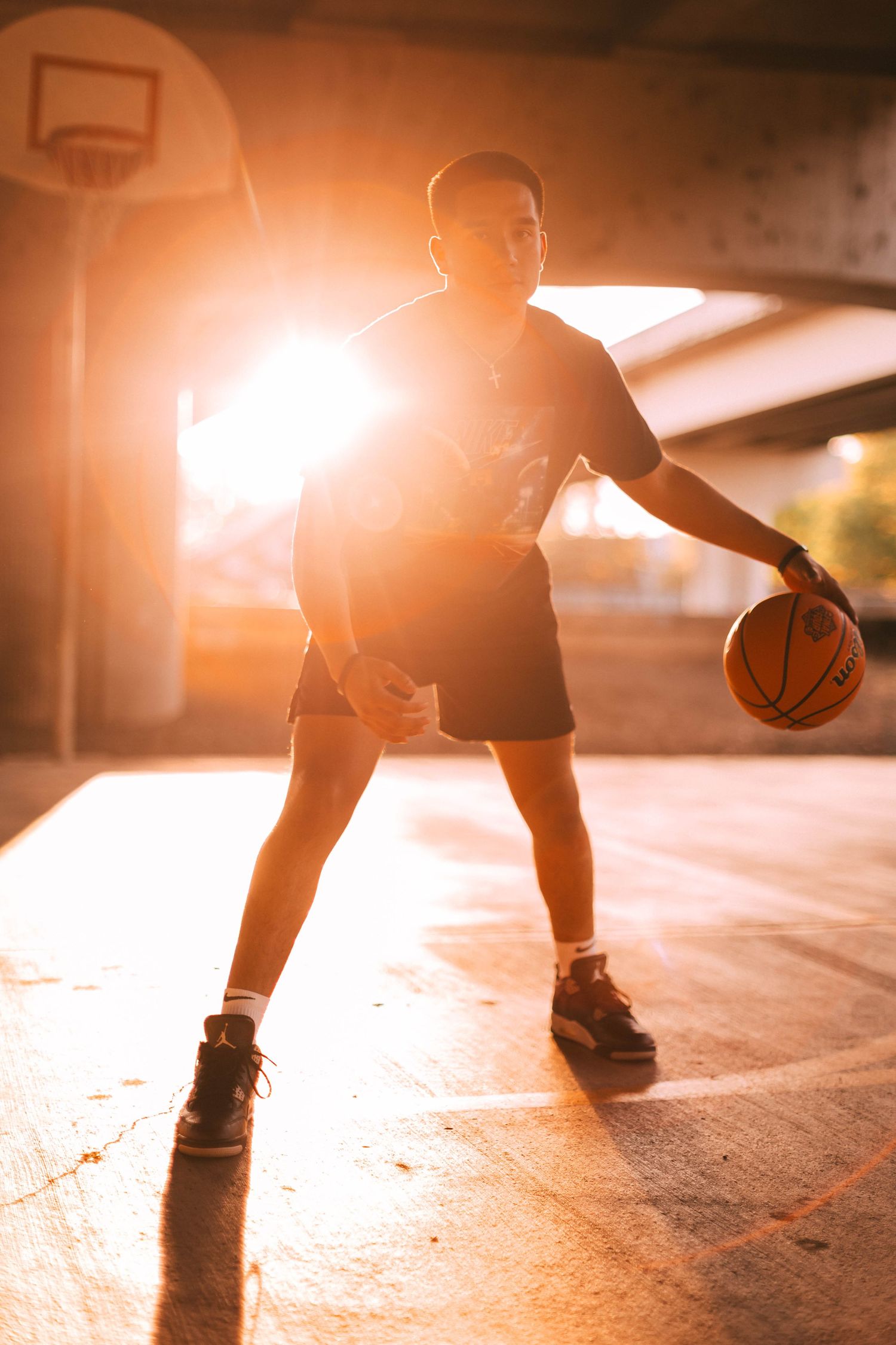 Silhouetted basketball player dribbling in a sunlit indoor court with dramatic shadows and warm golden lighting.