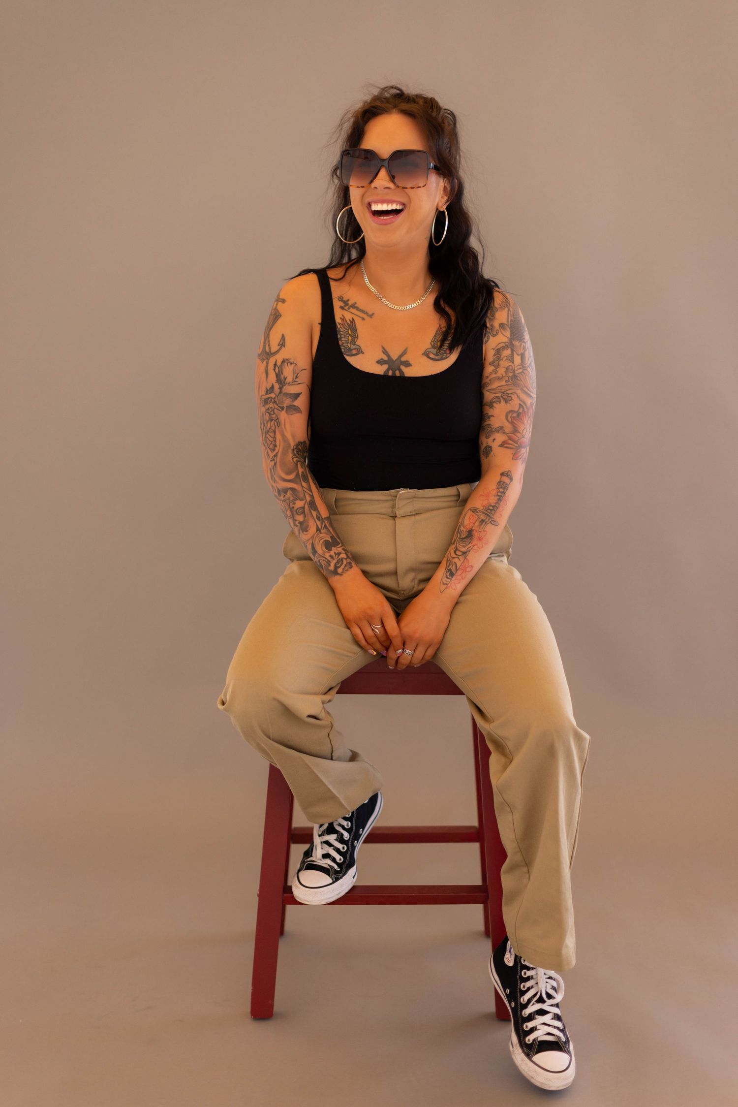 A person in khaki pants and black top poses playfully on a red chair against a beige backdrop wearing sunglasses.