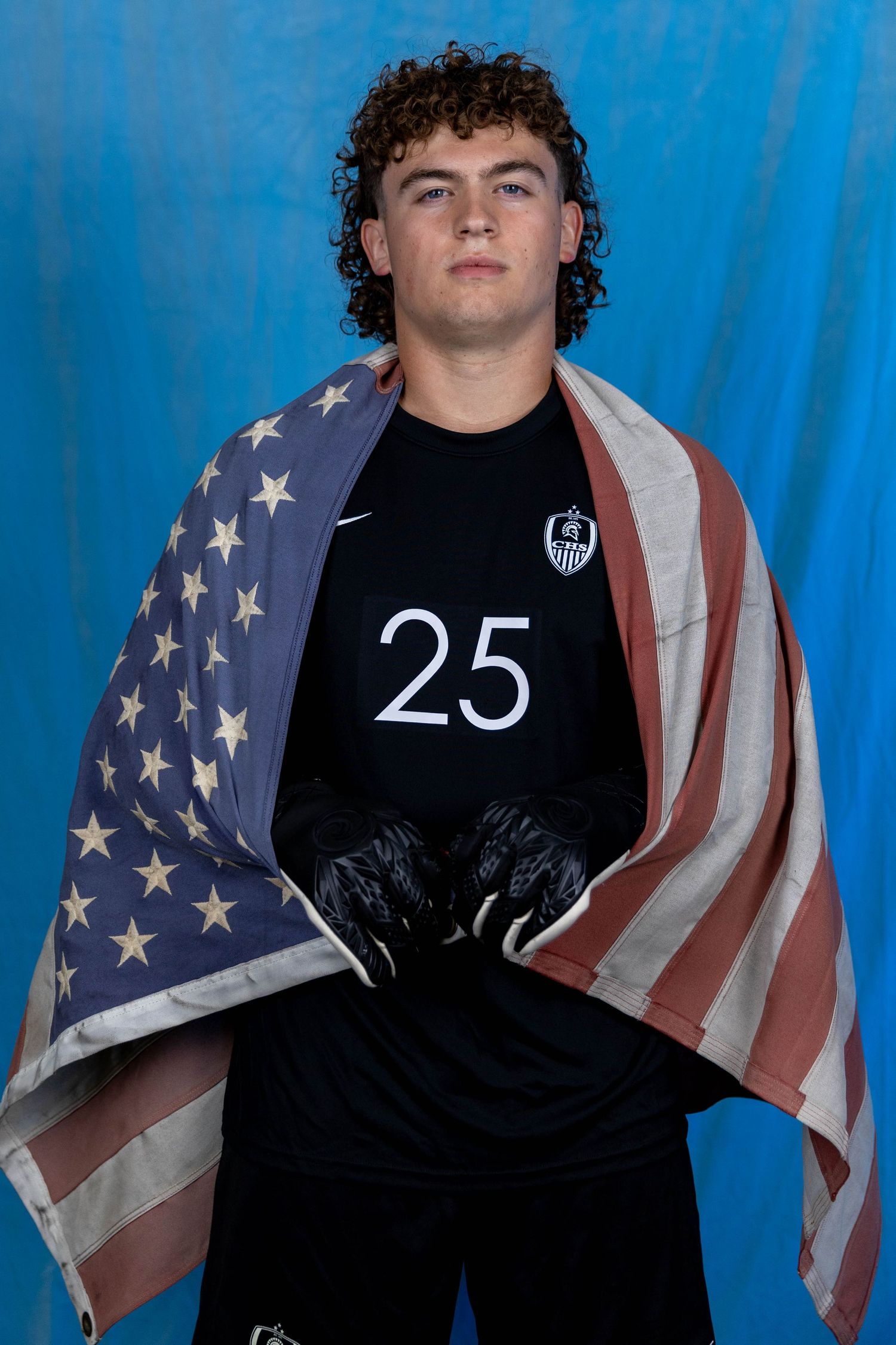 Athlete in black jersey number 25 poses with American flag draped over shoulders against blue backdrop.