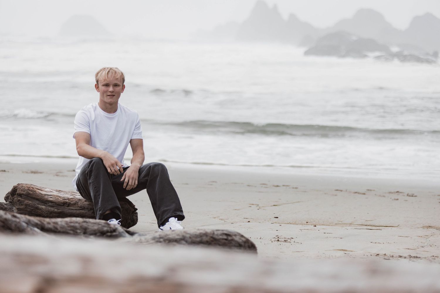 A person sits casually on driftwood at a misty beach during overcast weather.