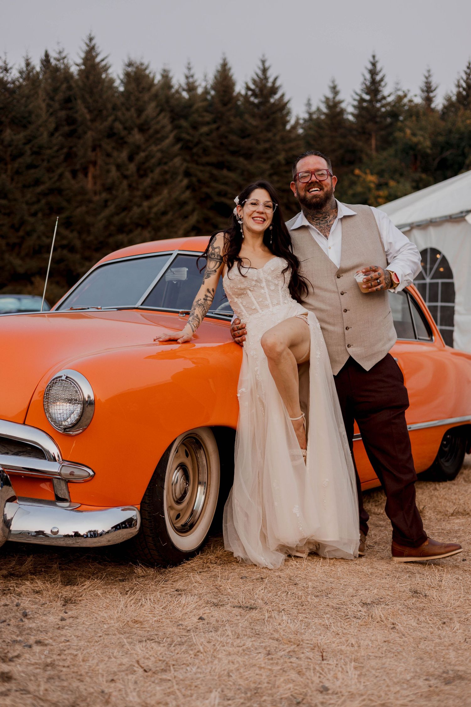 A vintage orange car serves as backdrop for romantic wedding photos with evergreen trees in the background.