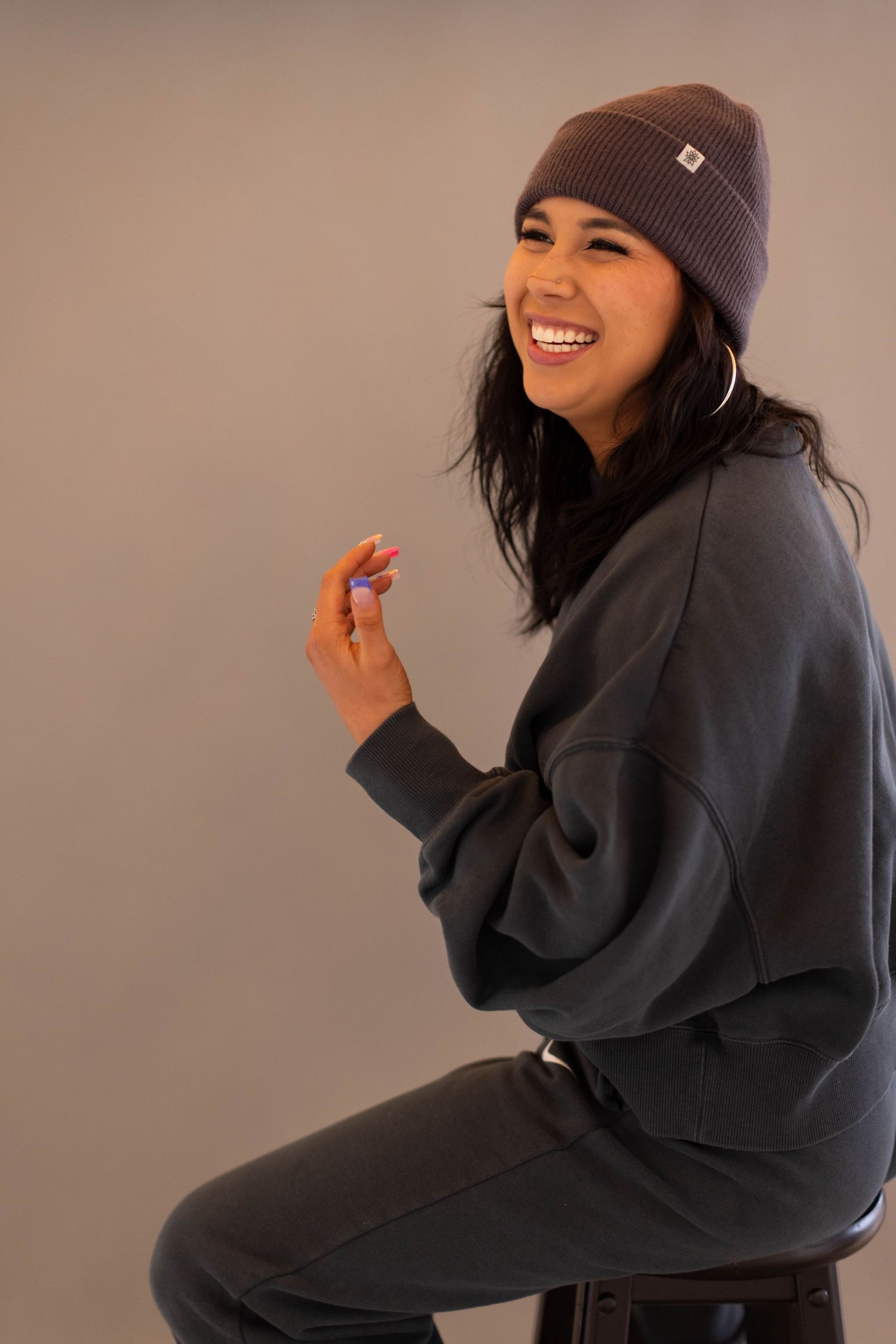 Person wearing a gray beanie and black sweater sits casually on a stool while smiling and posing for photos.