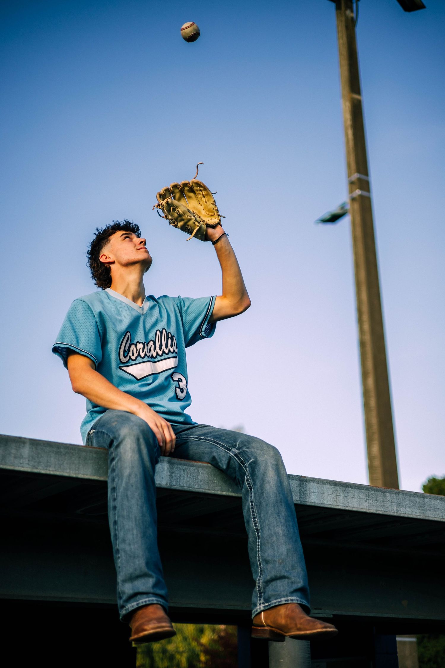 Baseball player in teal jersey sits on dugout catching ball against blue sky.