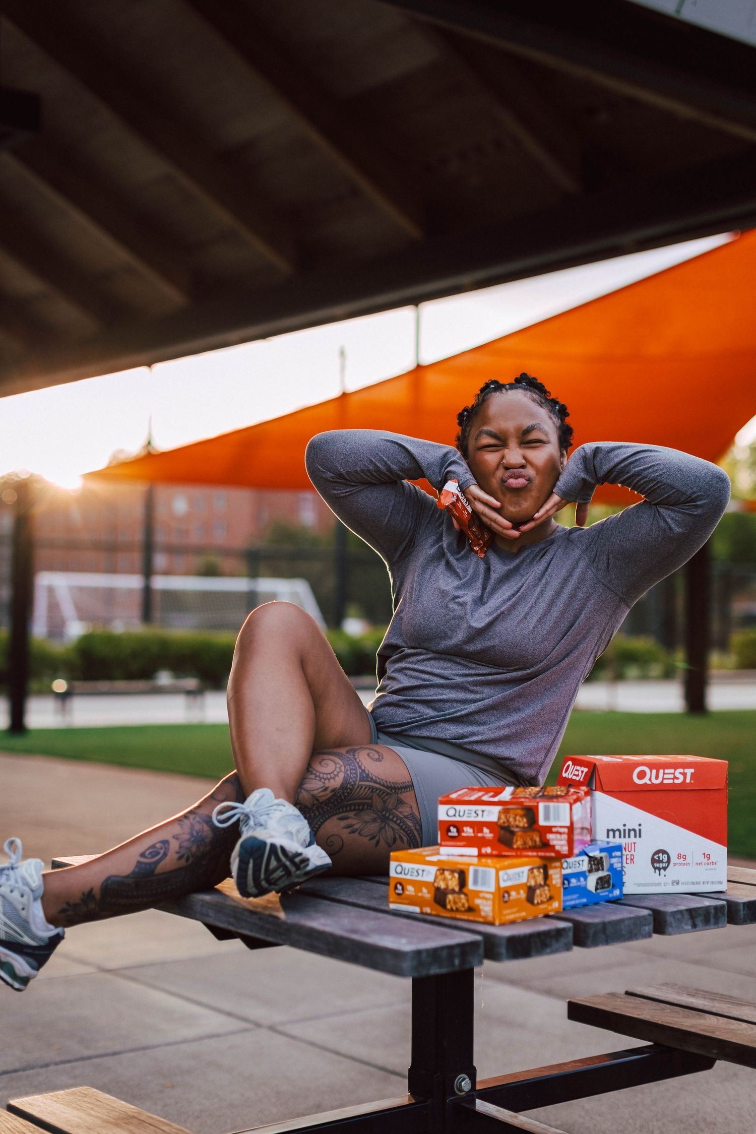 Person relaxing on outdoor bench with boxes of Quest protein bars spread out on table during sunset.