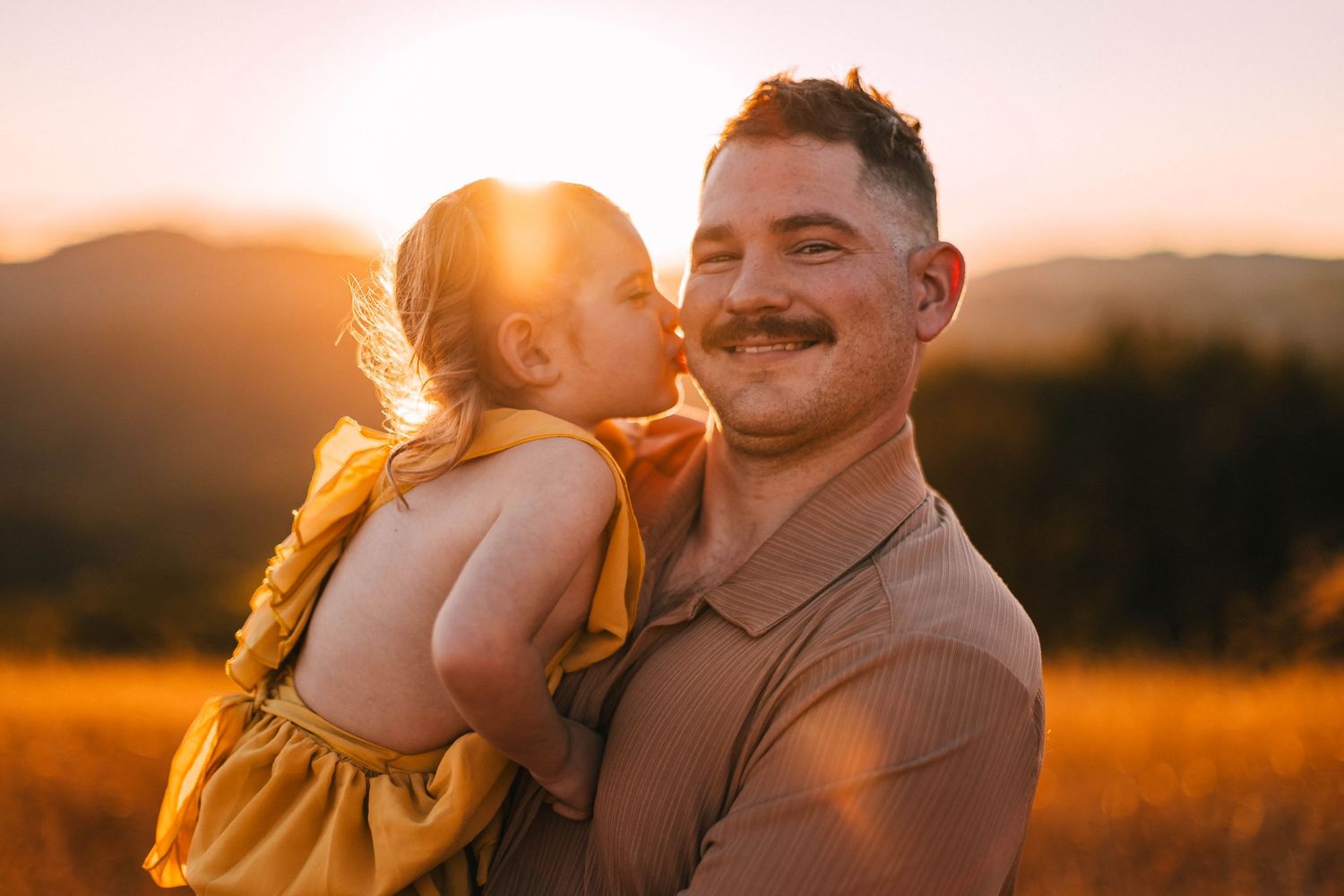 Couple embraces in golden sunlit field during sunset photoshoot creating romantic silhouettes against orange sky.