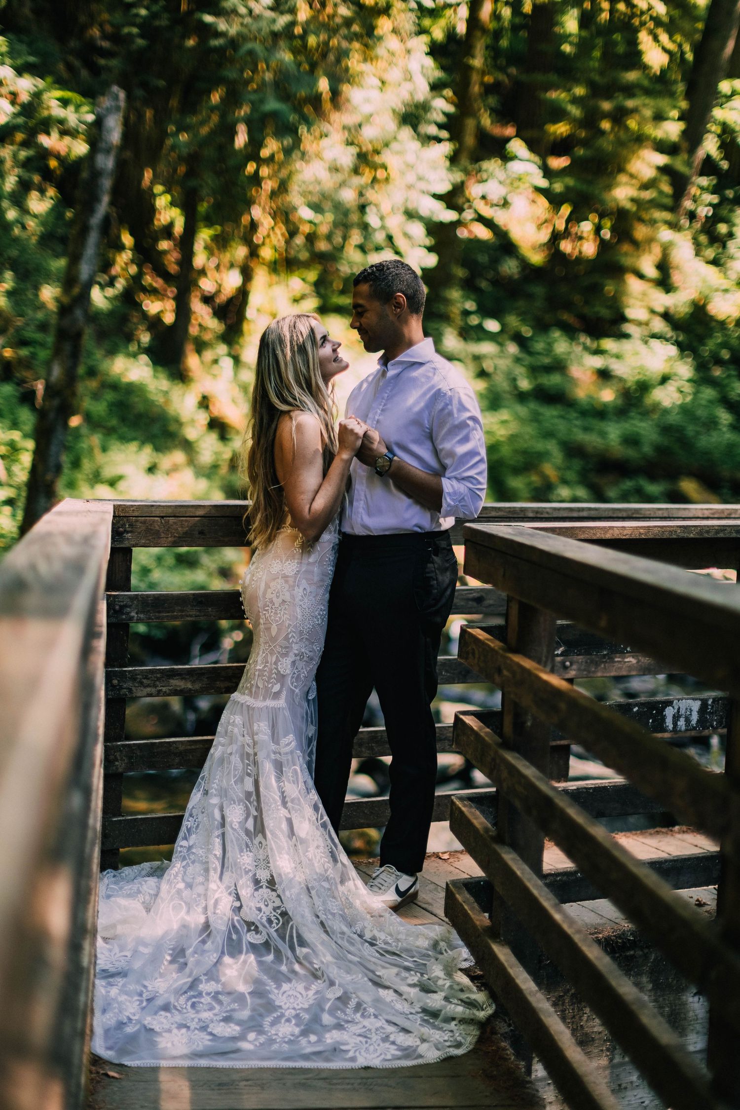 Couple in wedding attire sharing tender moments on a rustic wooden bridge in a forest setting.