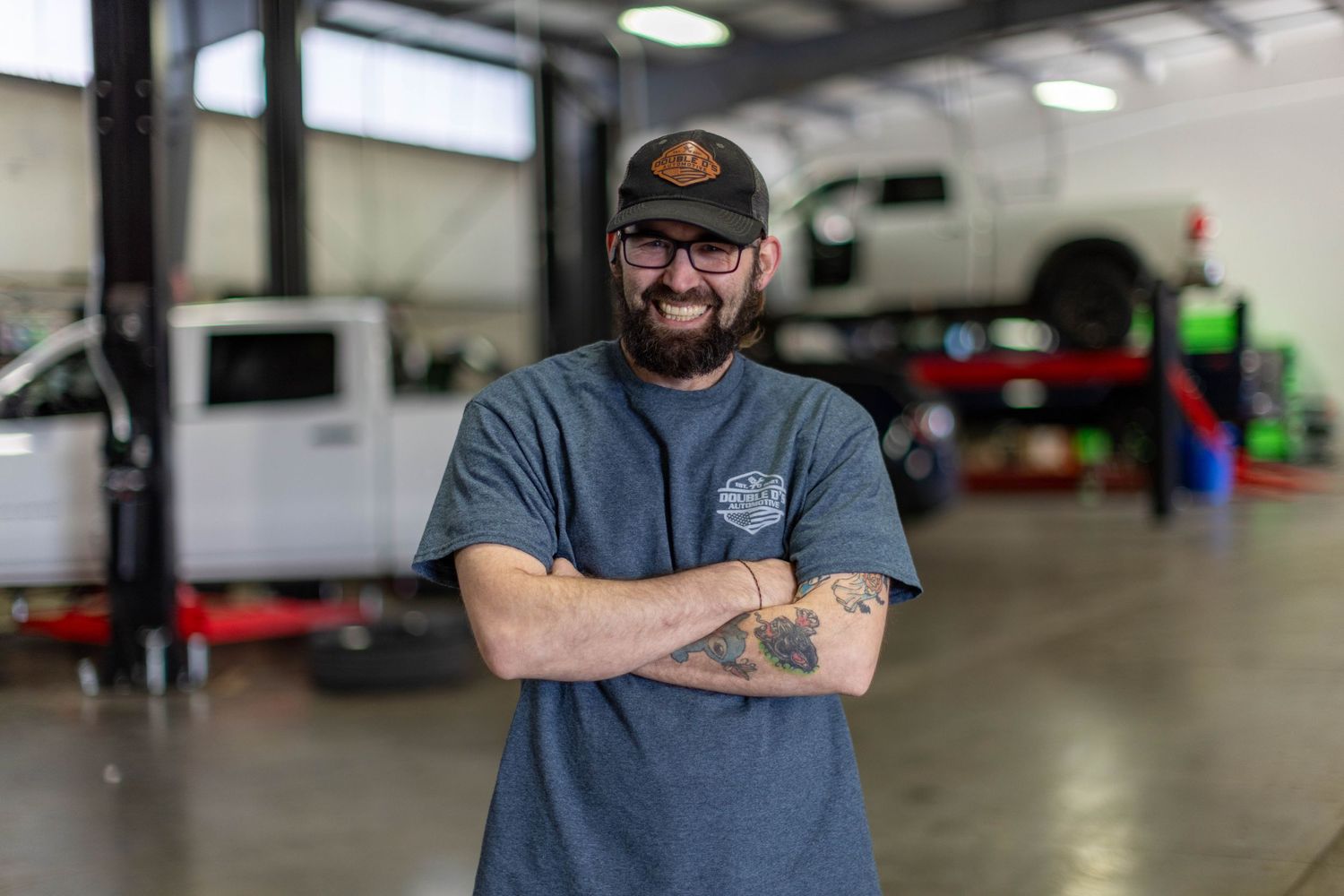 Auto mechanic wearing glasses and grey t-shirt stands confidently in repair shop with arms crossed.