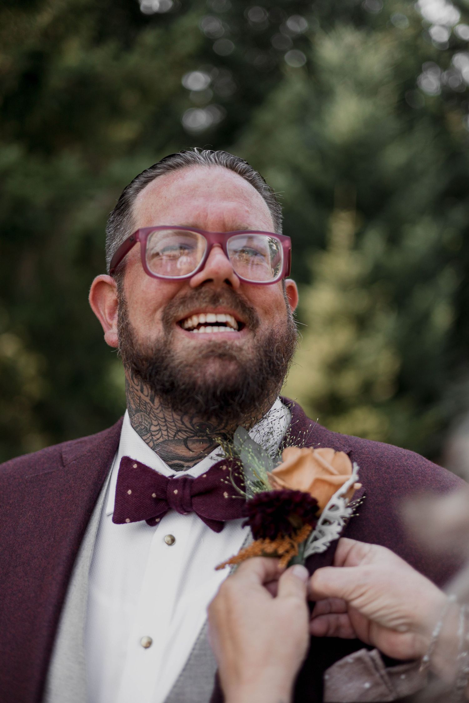 A person in a tuxedo and bow tie laughs joyfully while someone adjusts their burgundy boutonniere.