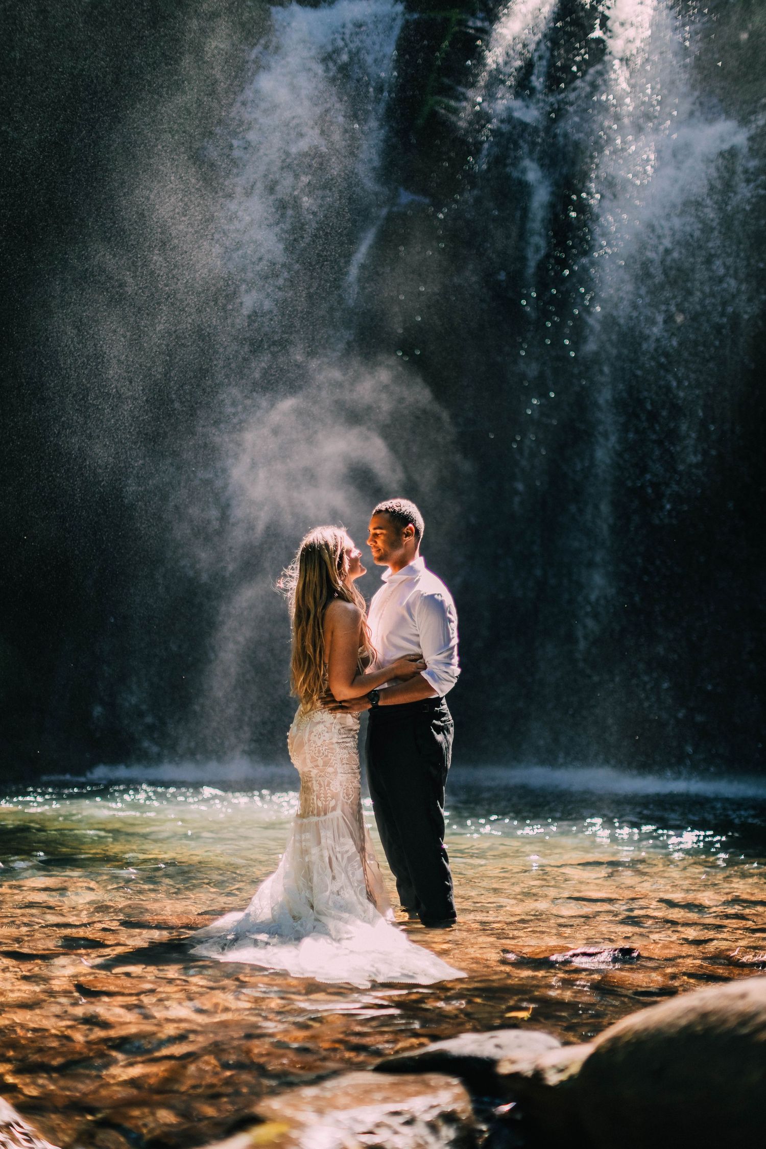 Newlyweds embrace by majestic waterfall with sunlight creating magical mist effect.