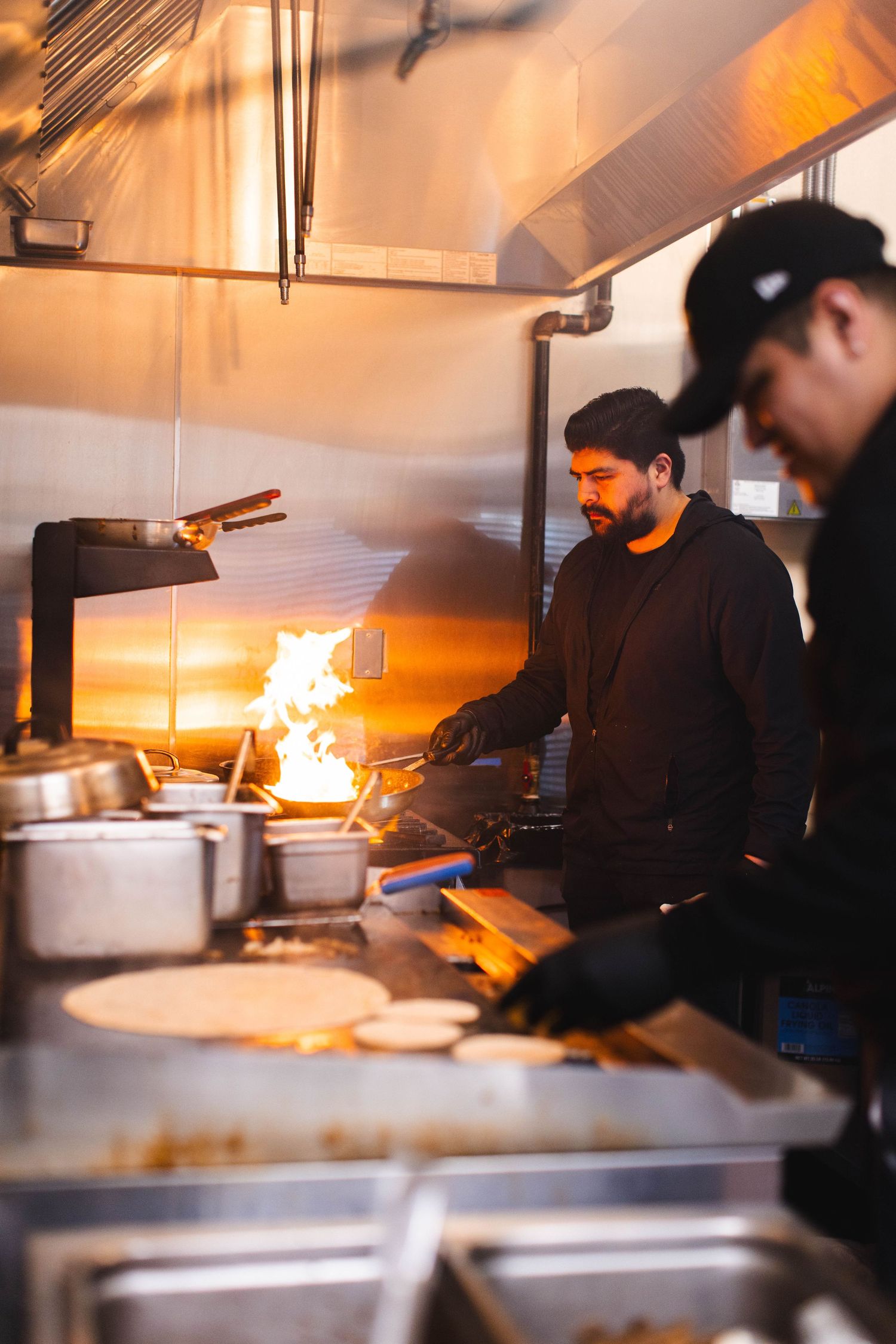 Chefs work in a busy restaurant kitchen with flames rising from the stovetop as they prepare meals during service.