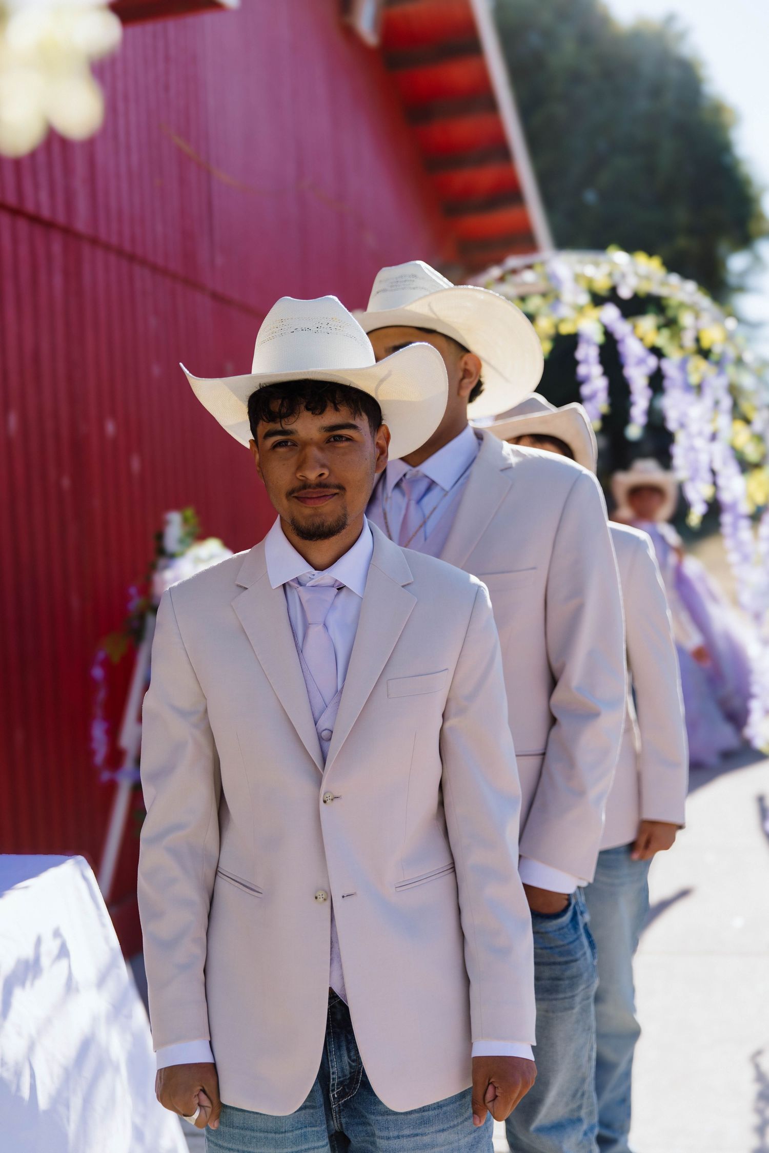 Groomsmen in light beige suits and white cowboy hats stand in line against a red barn wall at a western-themed wedding.