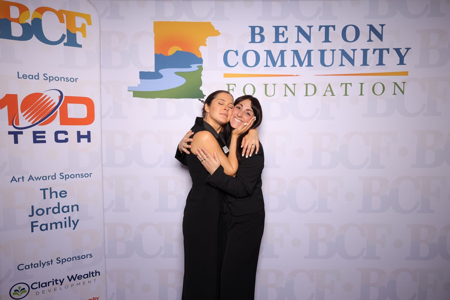 Two people pose playfully in front of a wall displaying corporate sponsor logos and banners at a community event.
