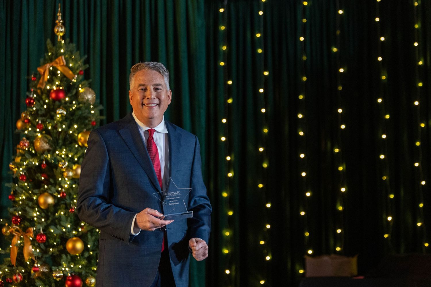 Someone in a navy suit stands by a decorated Christmas tree against a backdrop of twinkling holiday lights.