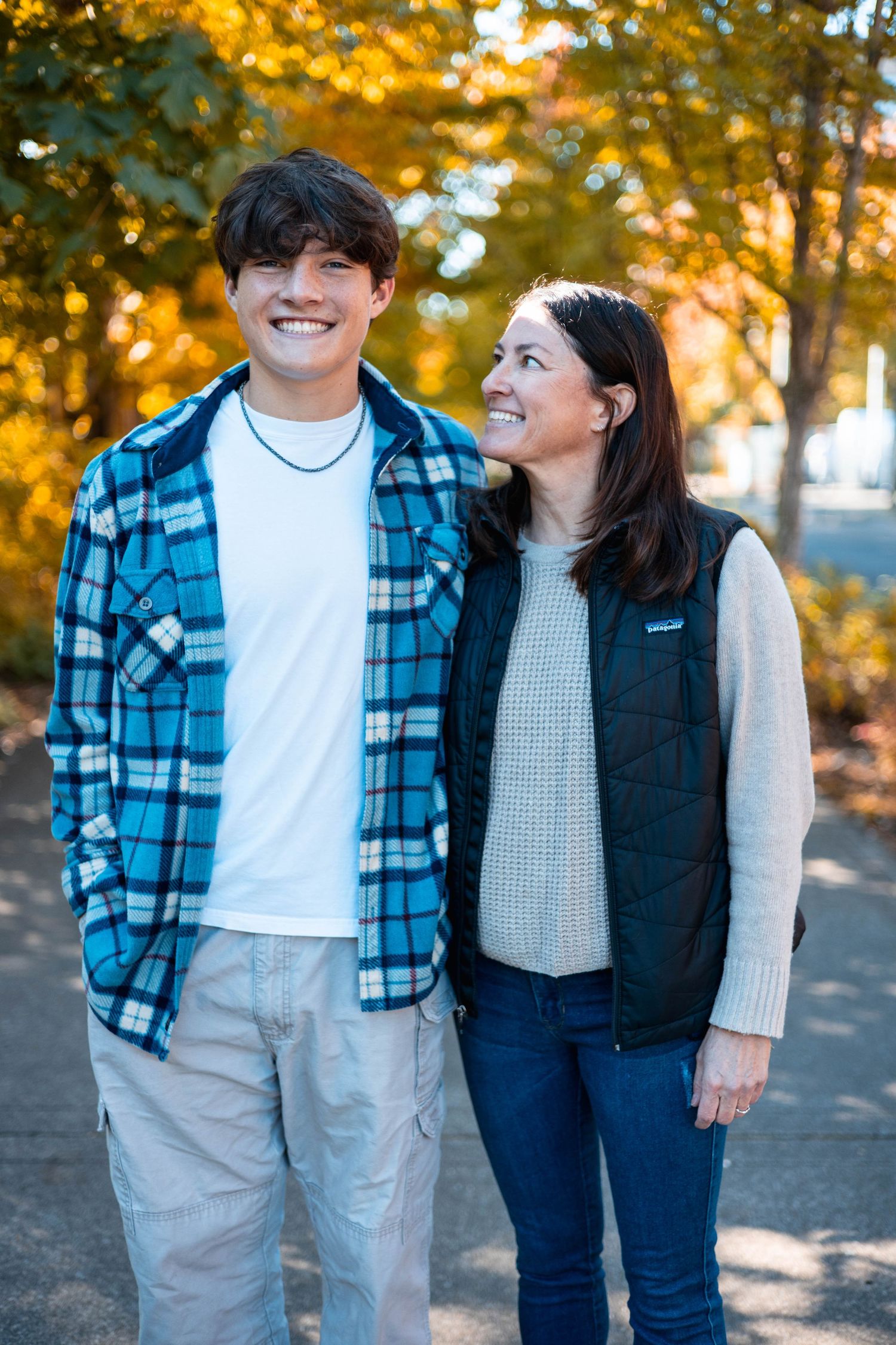 A couple poses together outdoors in autumn with yellow foliage in the background wearing casual clothing.