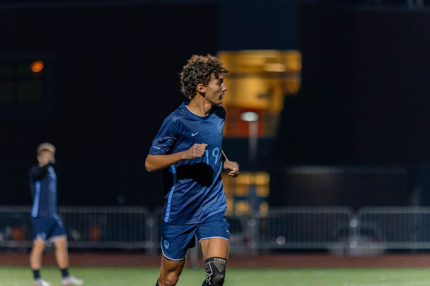 Soccer player in blue uniform dribbles ball on field during nighttime match under stadium lights.