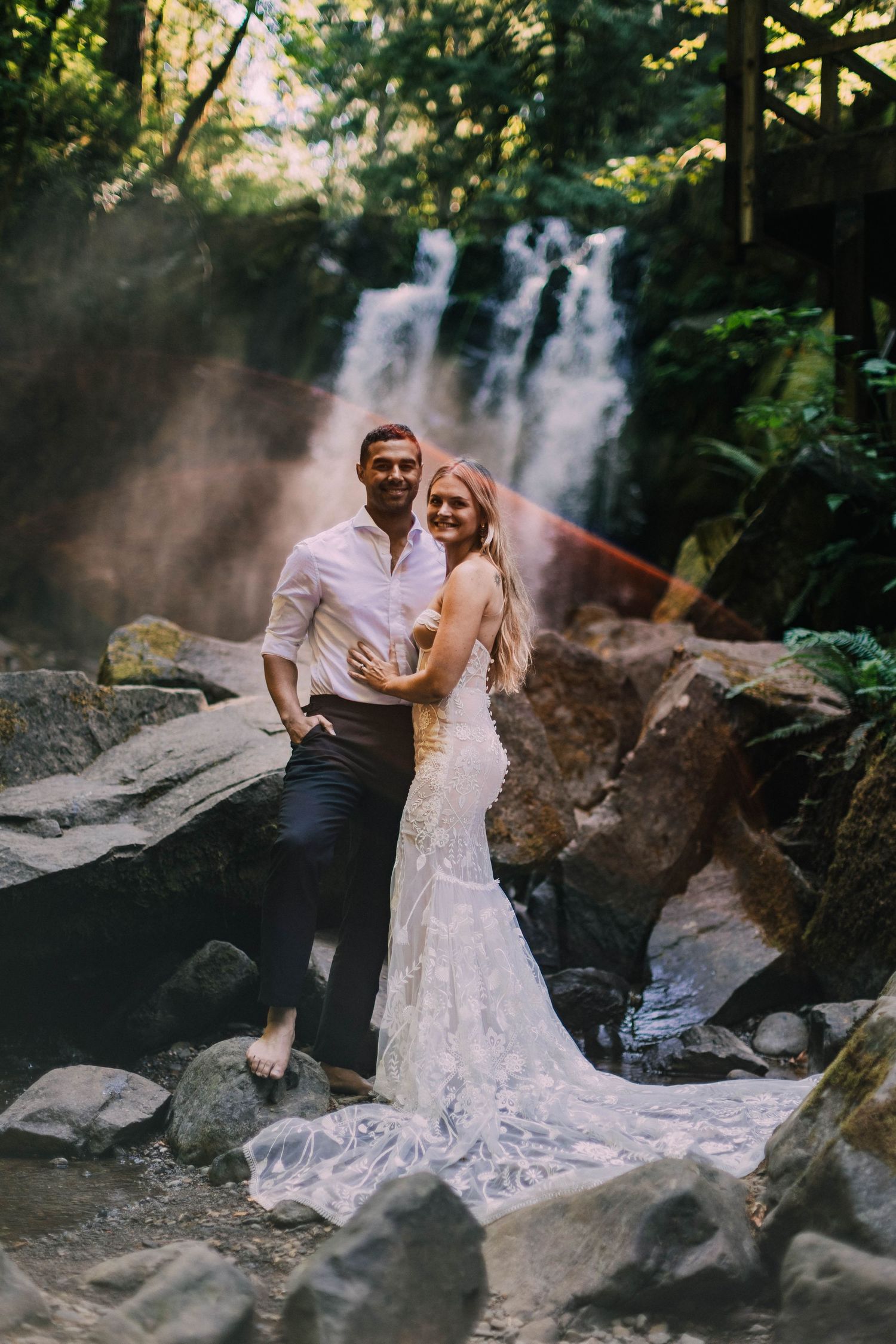 Wedding couple poses near misty waterfall on rocky terrain during outdoor photo session.
