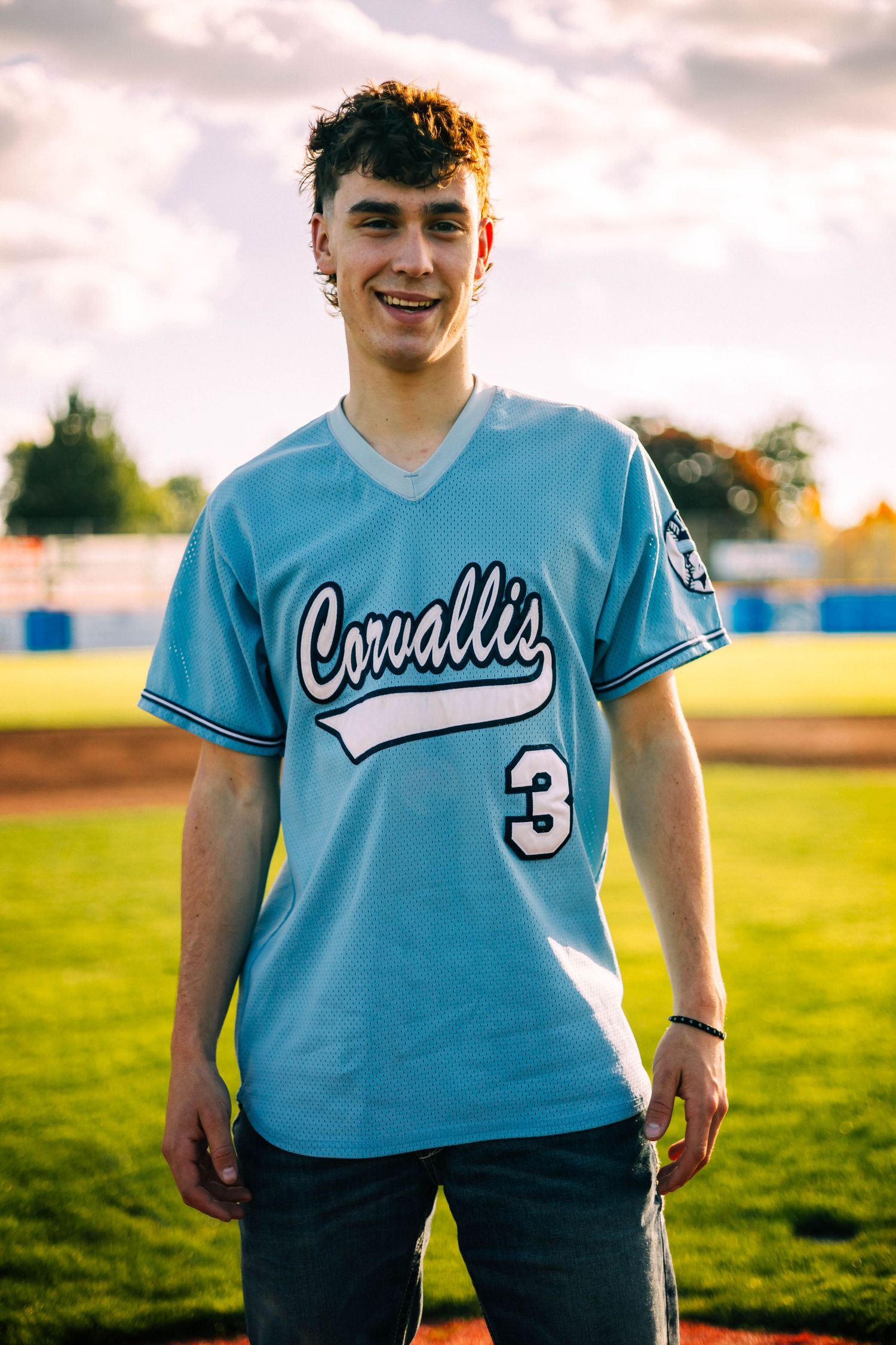 Baseball player in light blue Corvallis team jersey stands in sunny field at golden hour.