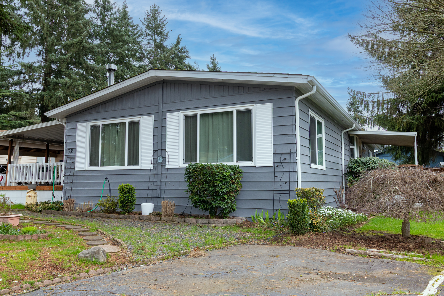 Gray single-story ranch home with attached carport and evergreen trees in the background under blue skies.