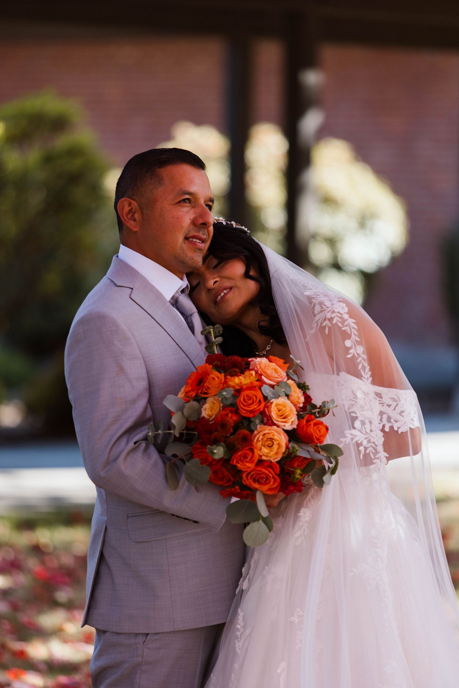 A wedding couple embraces outdoors while holding a vibrant orange and red rose bouquet.