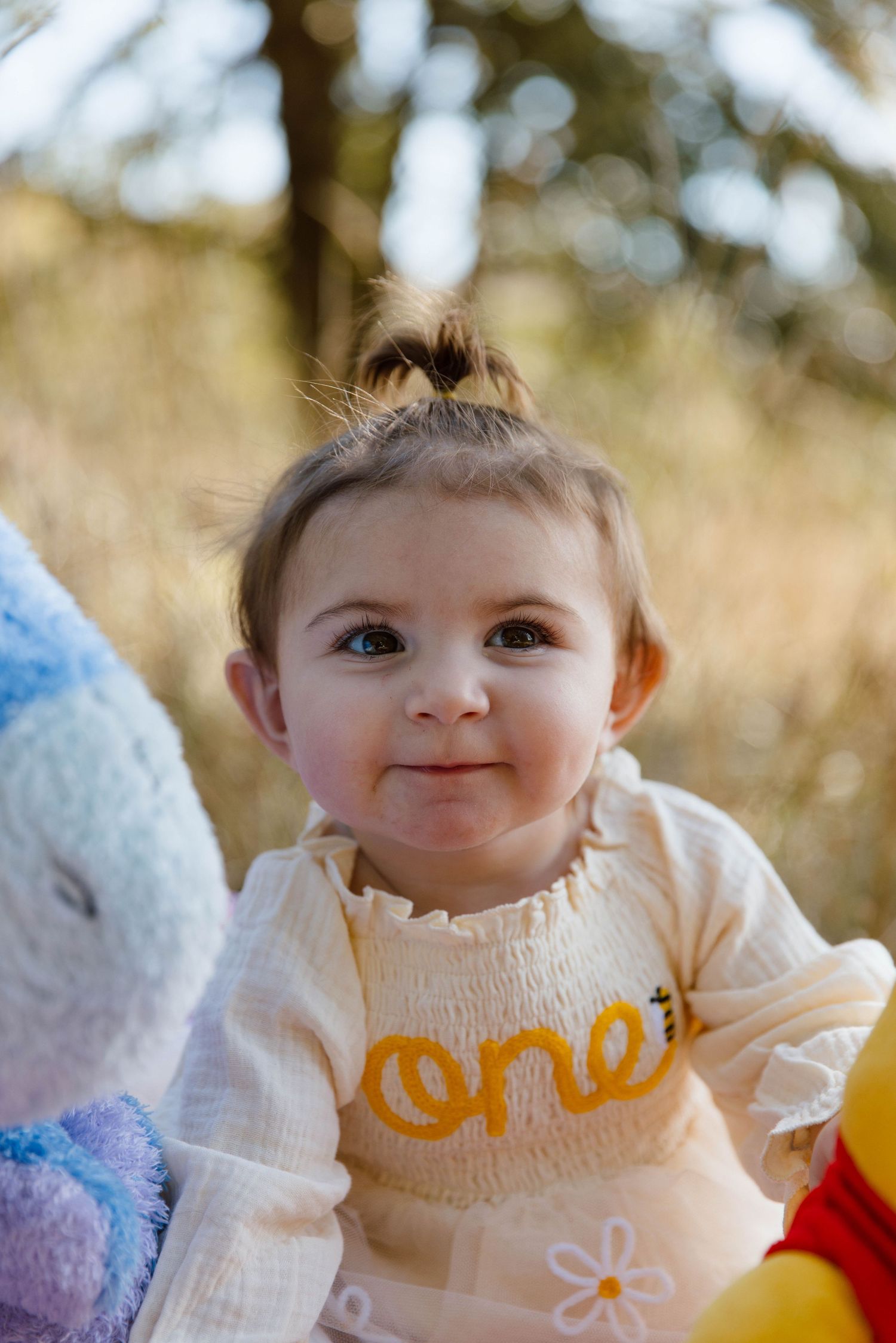 A baby wearing a cream-colored sweater smiles while sitting outdoors on a sunny day.