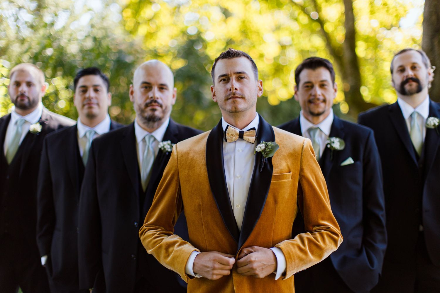 A group of groomsmen in formal attire stand together with two wearing golden jackets at an outdoor wedding.