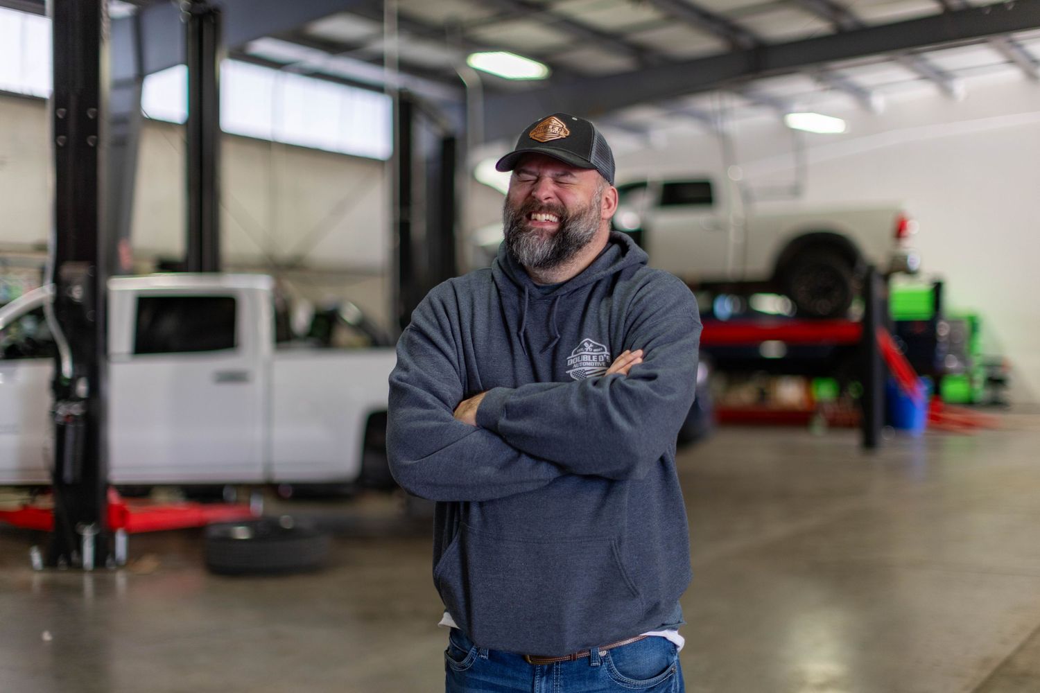 Auto mechanic in a gray sweatshirt stands with arms crossed in a repair shop garage with vehicles in background.