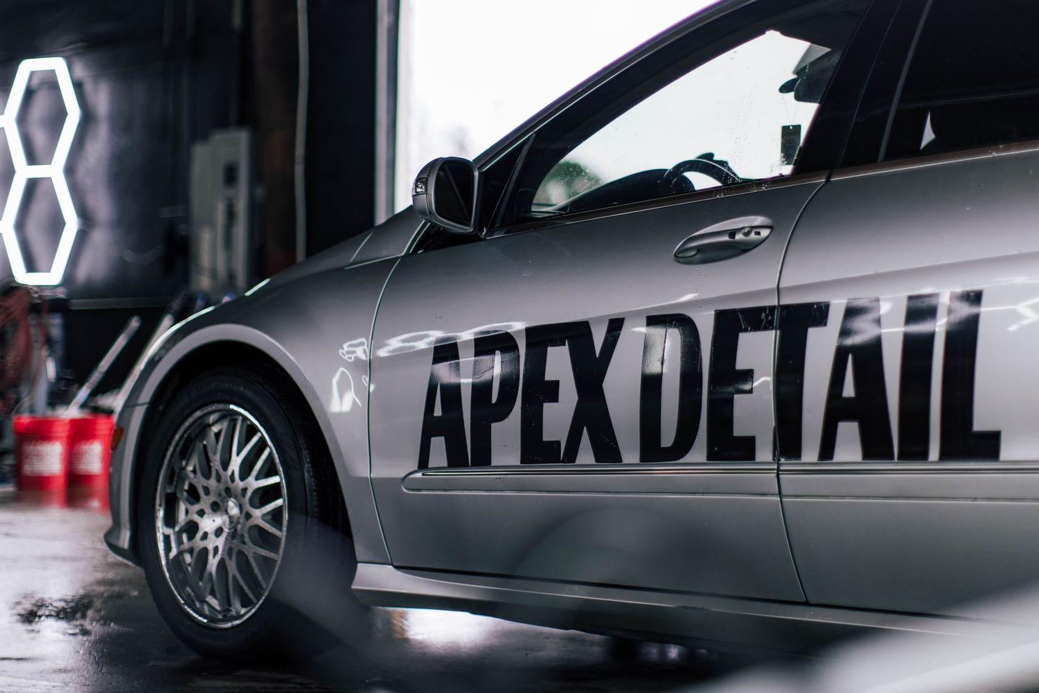A silver car with APEX DETAIL branding on the side panel sits in a garage with neon lighting in the background.
