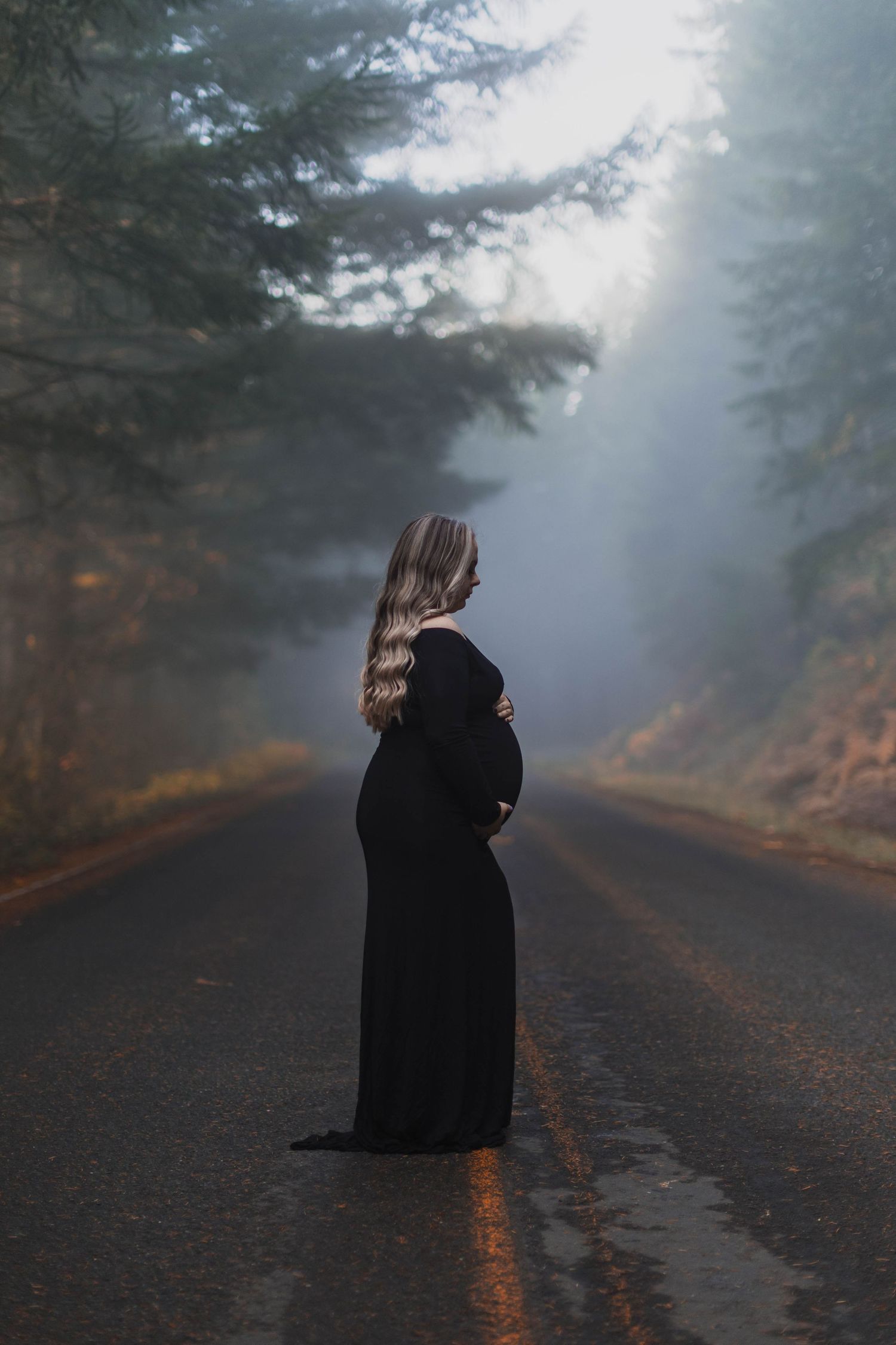 A silhouetted figure in a black coat stands on a misty forest road during twilight.