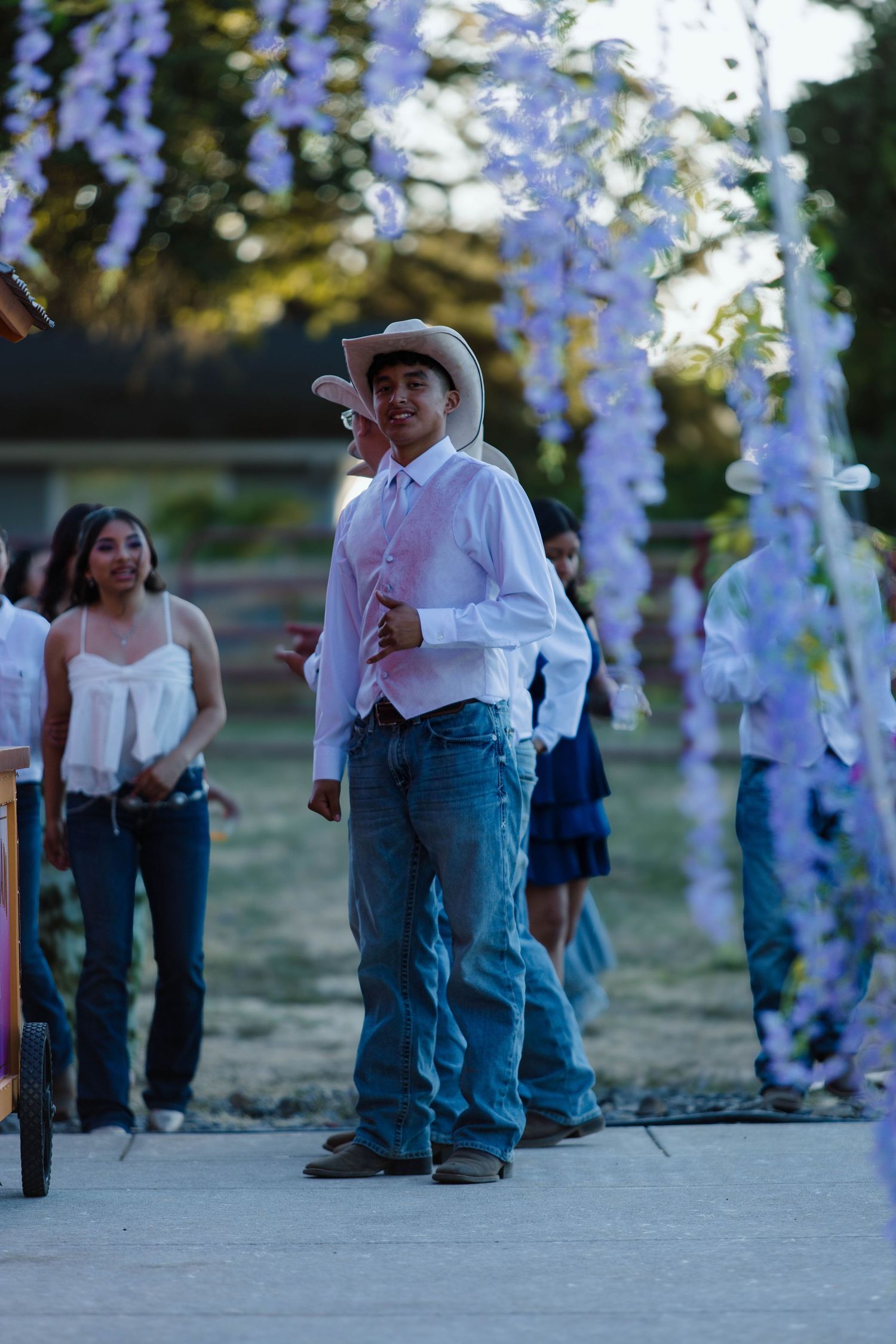 Guests in cowboy boots and jeans gather beneath hanging purple wisteria decorations at an outdoor event.