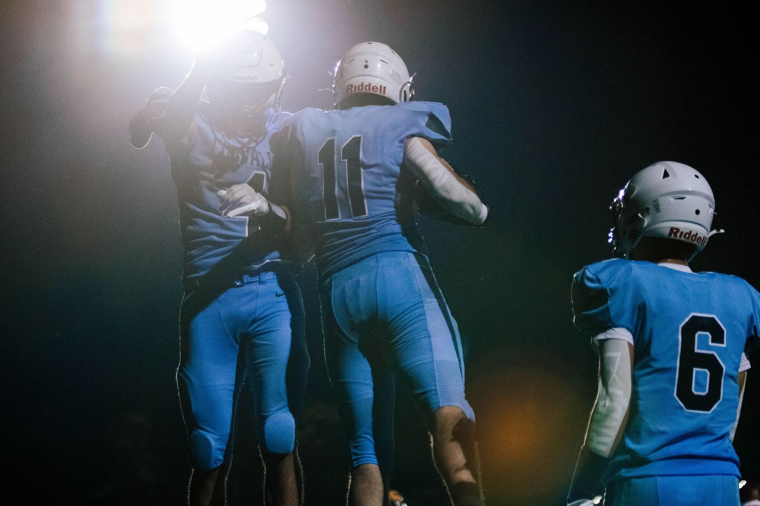 American football players in blue jerseys and white helmets huddle together on a dark field during a game.