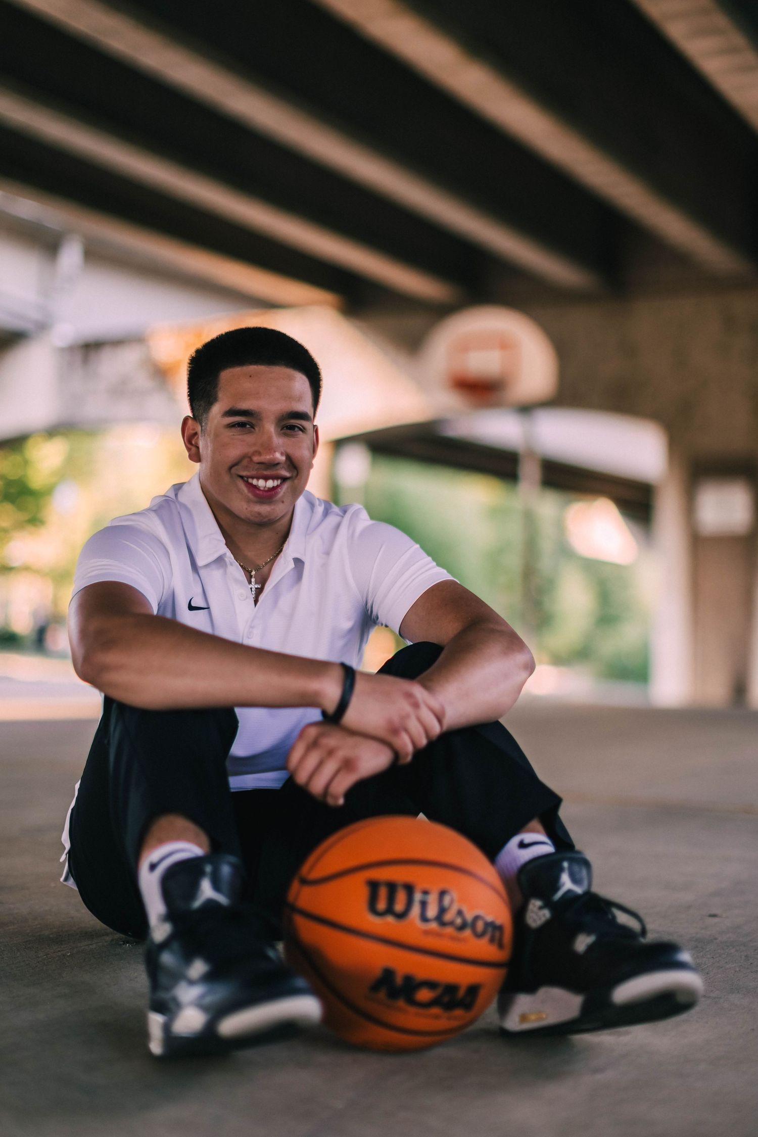 Young athlete in white polo shirt sits with Wilson NCAA basketball under covered outdoor area.