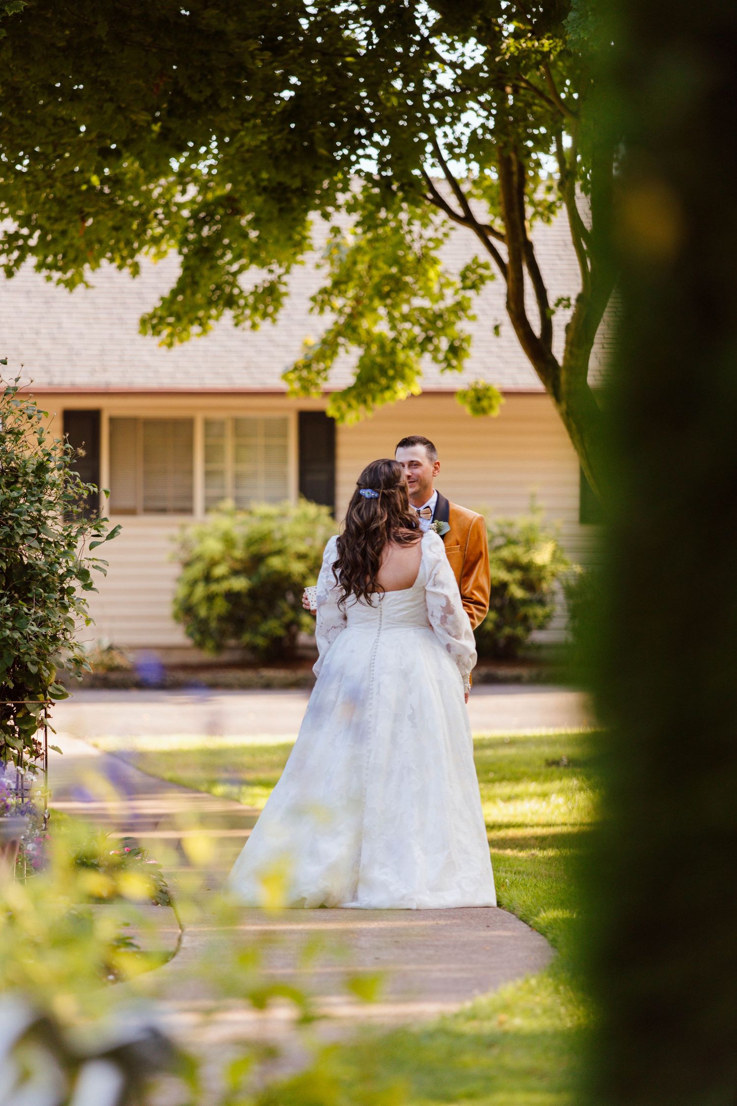 A couple in wedding attire shares a romantic moment in a sunlit garden setting with trees framing the scene.