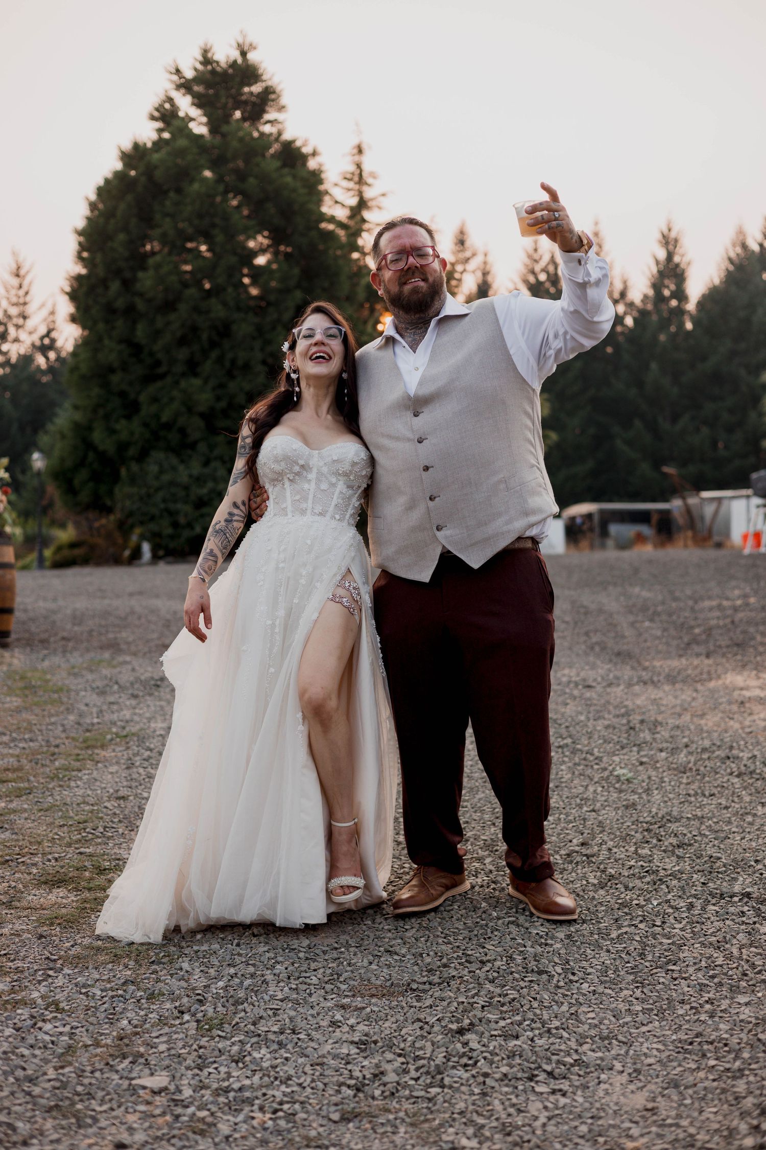 Wedding couple poses together on a gravel path with evergreen trees in the background at sunset.