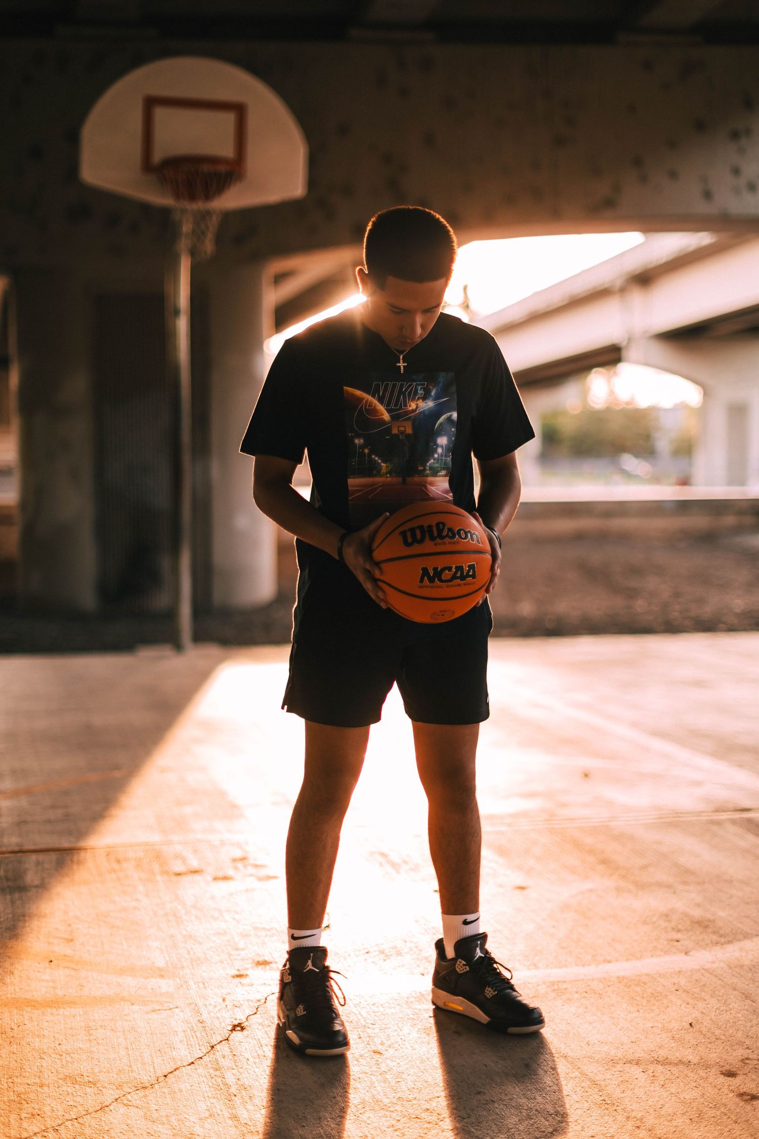 Low angle view of a basketball player's legs and shoes dribbling a ball under a bridge at sunset.
